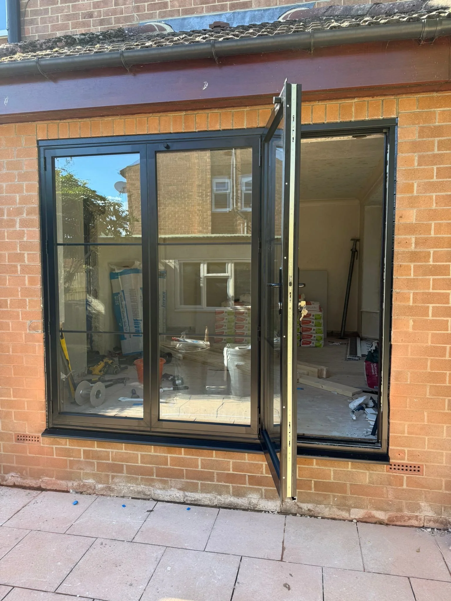 Under construction or renovation in progress at a house, featuring an installed black metal-framed sliding glass door with one panel open, inside is filled with construction tools and materials, such as a bucket, tools, and stacks of boxes, with a brick wall outside and a paved sidewalk in the foreground.