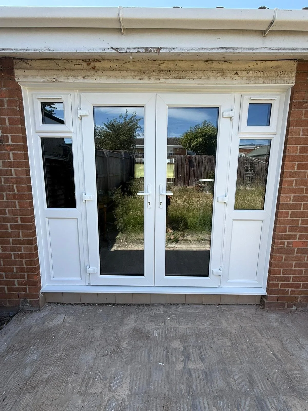 White double-glass patio doors with side panels, set in a brick wall, reflecting a backyard with trees, a fence, and a cloudy sky.