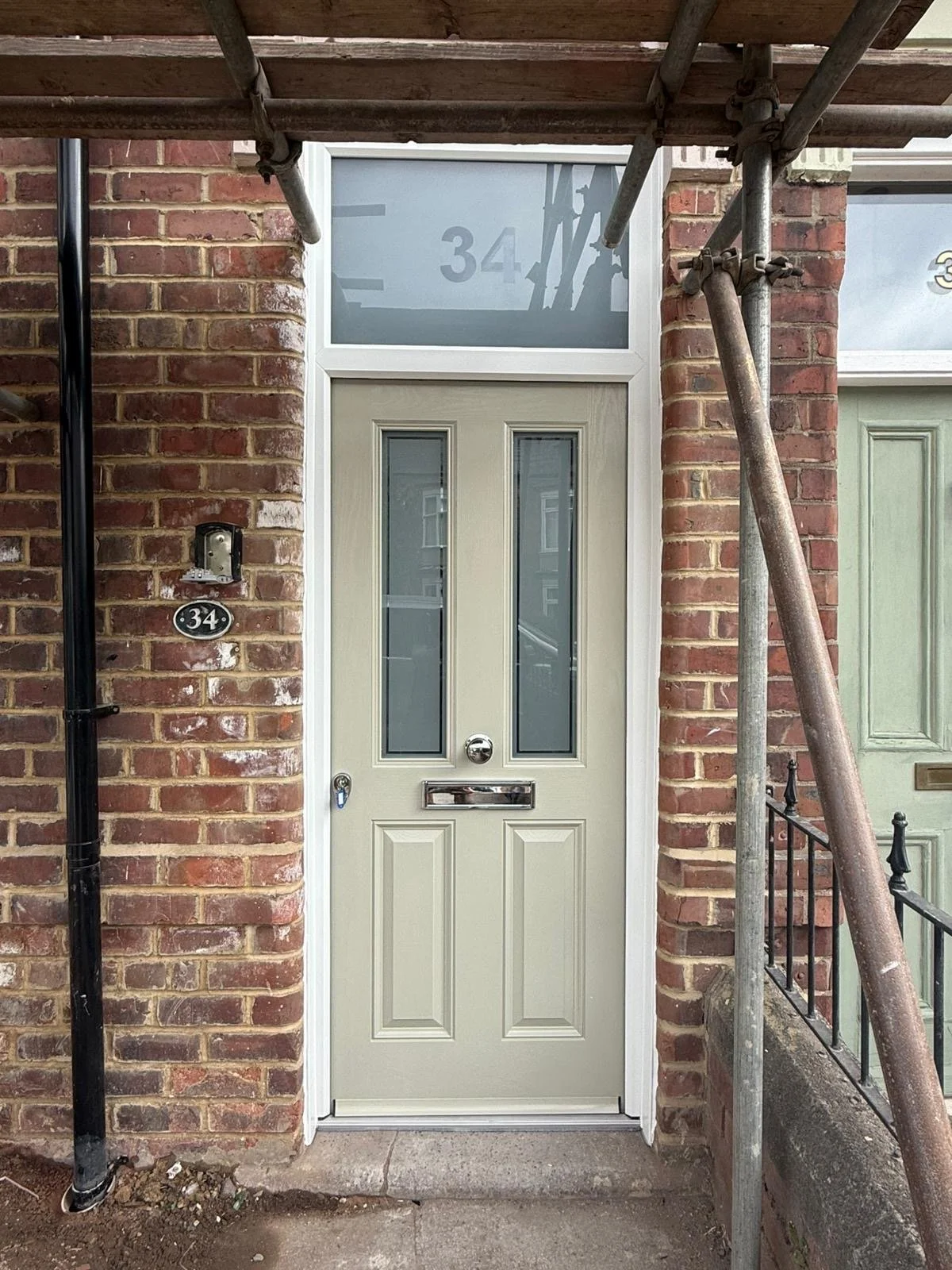 Front door of a brick building with a light-colored door, two narrow windows, a mail slot, and a round door knob, surrounded by scaffolding.