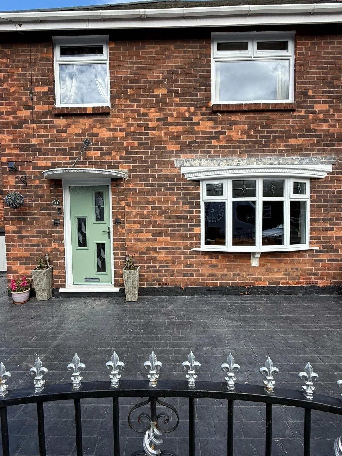 Front exterior of a brick house with a green door, white-framed bay window, two upper windows, and decorative flower pots on dark paved ground.