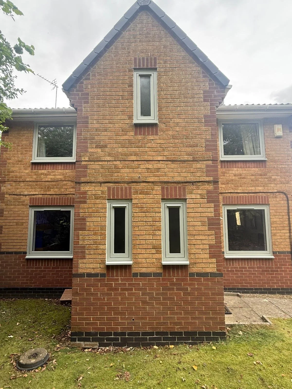 Front view of a brick house with six windows, three on each side, and a small triangle-shaped section at the top with a tall, narrow window. The upper part of the house is brick, and the lower part has a dark brick or stone base. There are trees on t