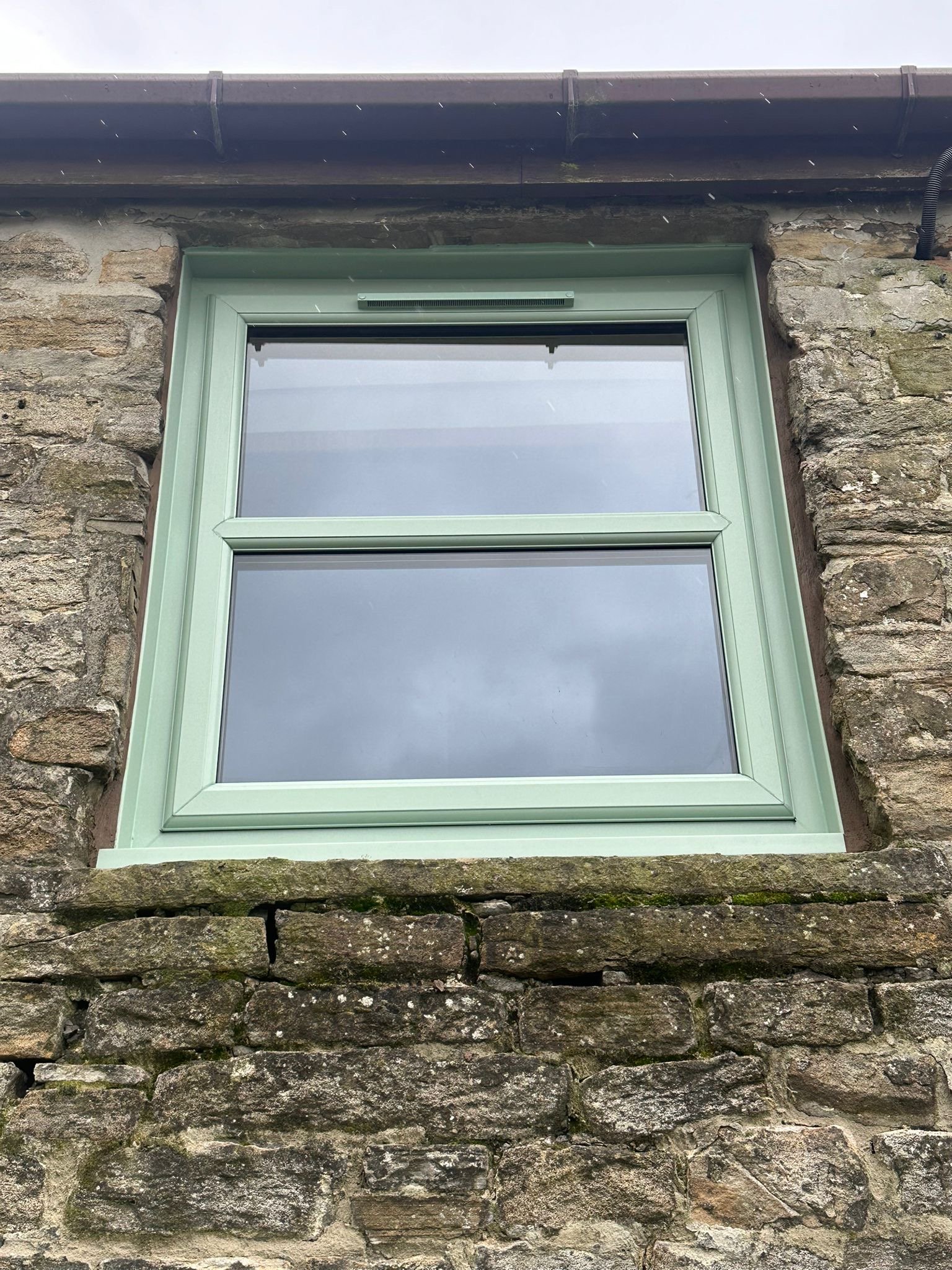 Close-up of a window with a green frame on an old stone building, showing the stone wall and part of the roof.