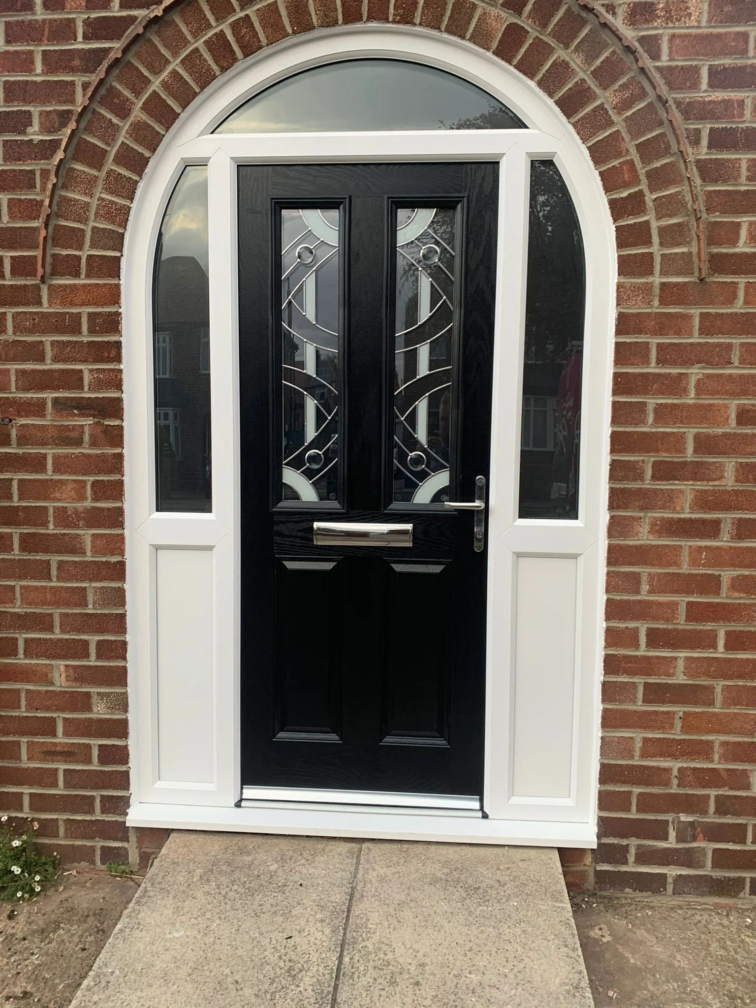 A black front door with decorative glass panels is set in a brick house facade. The door has a silver handle and letter slot. There is a white trim and an arched window above the door. A small concrete stoop leads to the door, with some soil and a sm