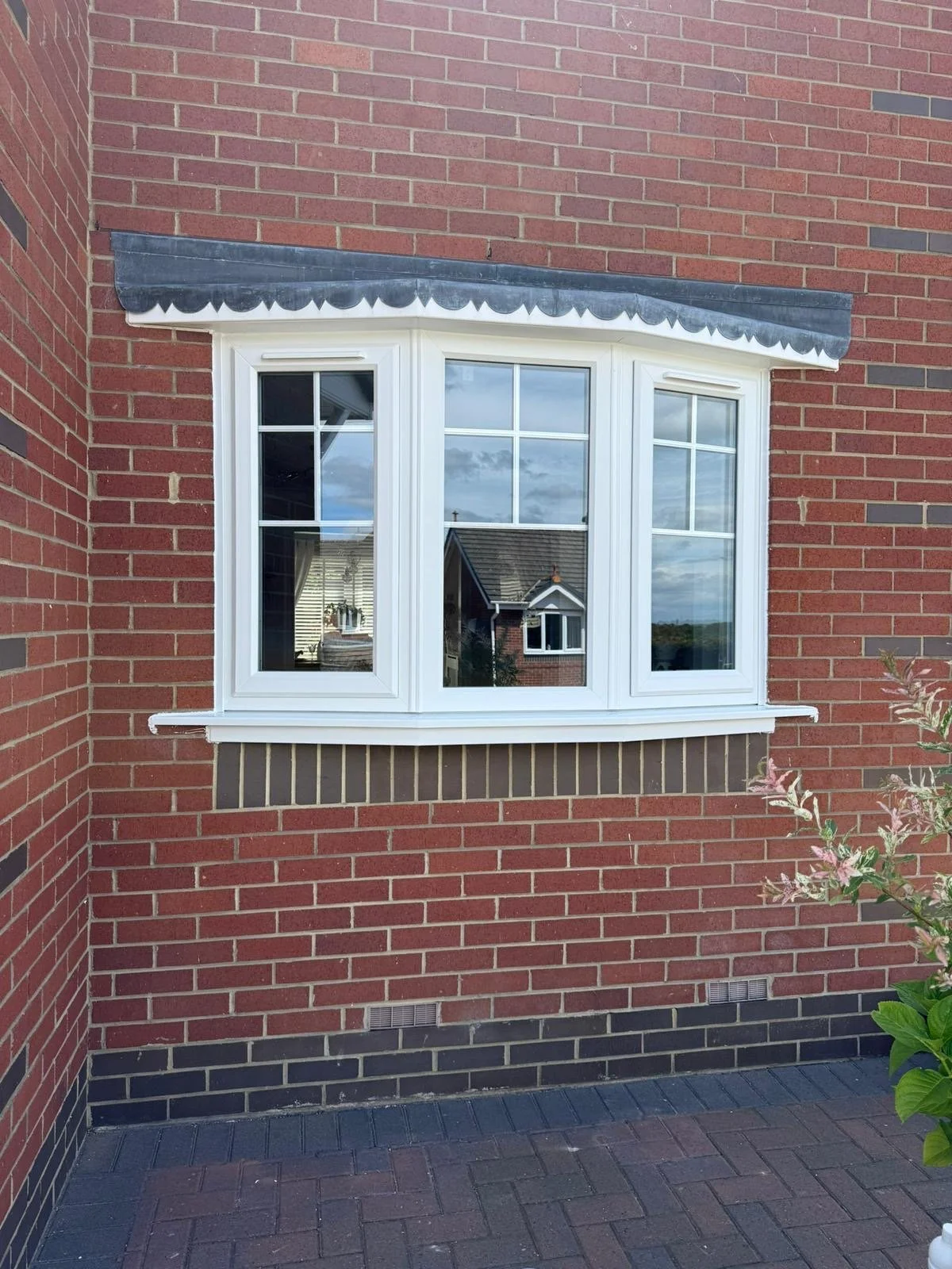 A large white bay window set into a red brick wall, with a decorative black awning above it and a brick sill below. The window reflects neighboring houses and the sky.