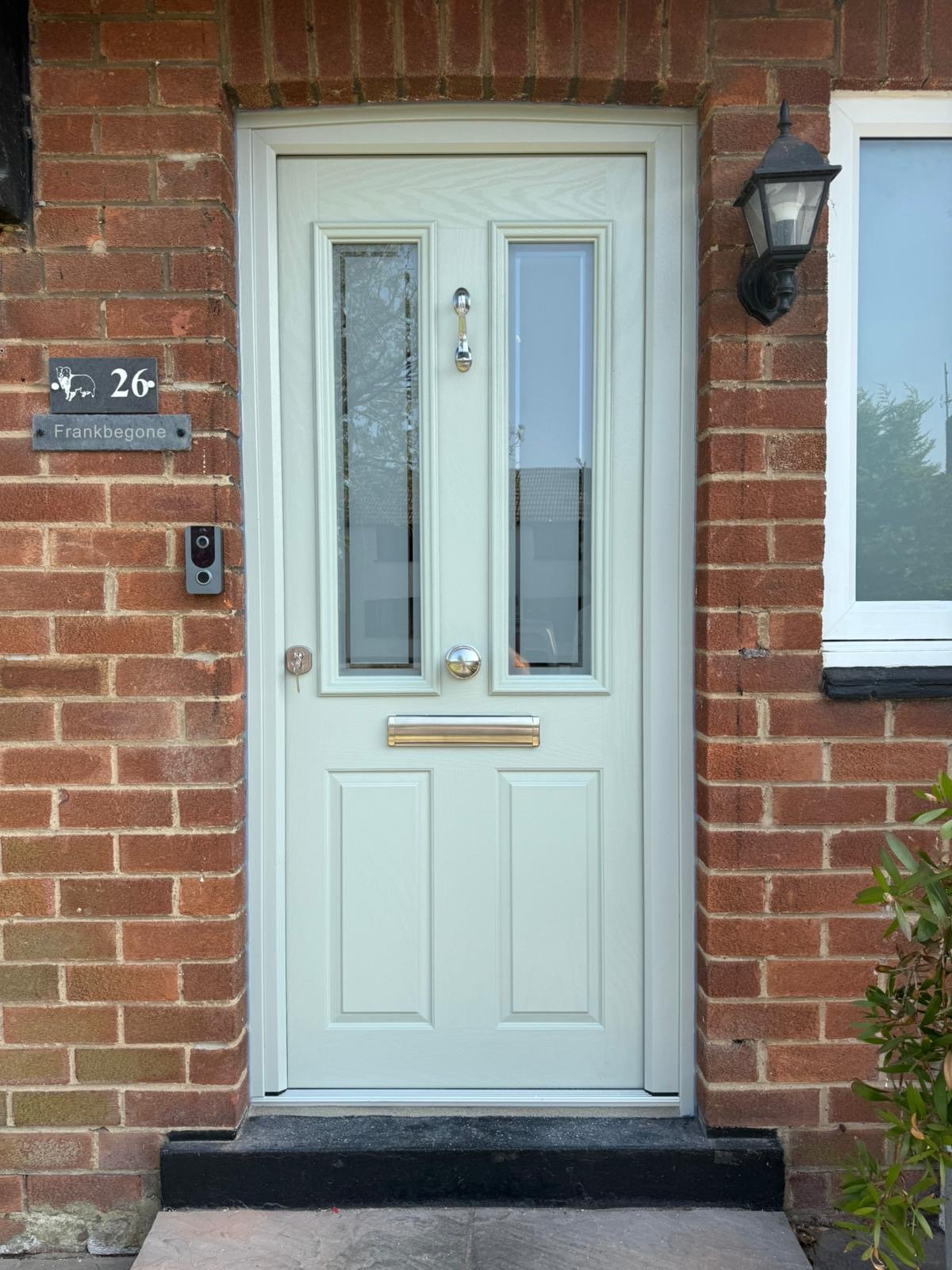 Front door of a brick house with a white door featuring glass panels, a mail slot, and a door knocker, flanked by a lantern-style light fixture and house number and name plaques.