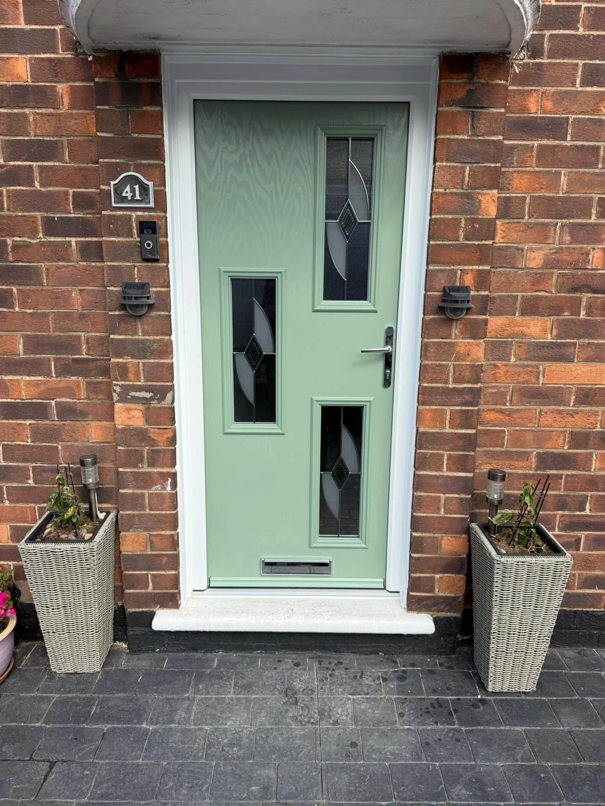 Light green front door with decorative glass panels on a brick house, flanked by two planters with plants, and brick wall background.