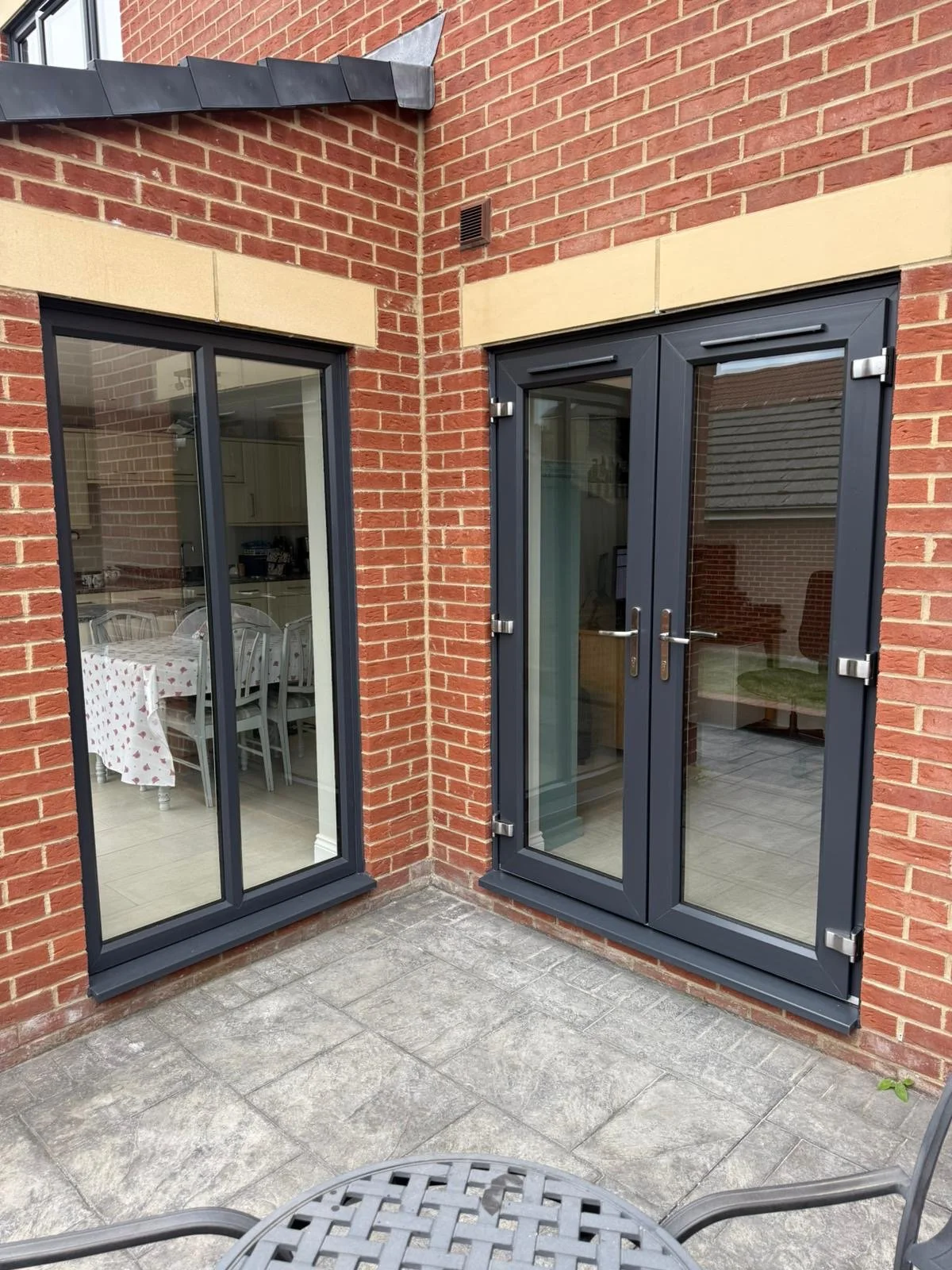 A brick house exterior with two large glass sliding doors, one door is double and the other is a single with a window. Inside, a dining table with a tablecloth and chairs is visible through the left door. The patio floor is made of gray tiles, and th
