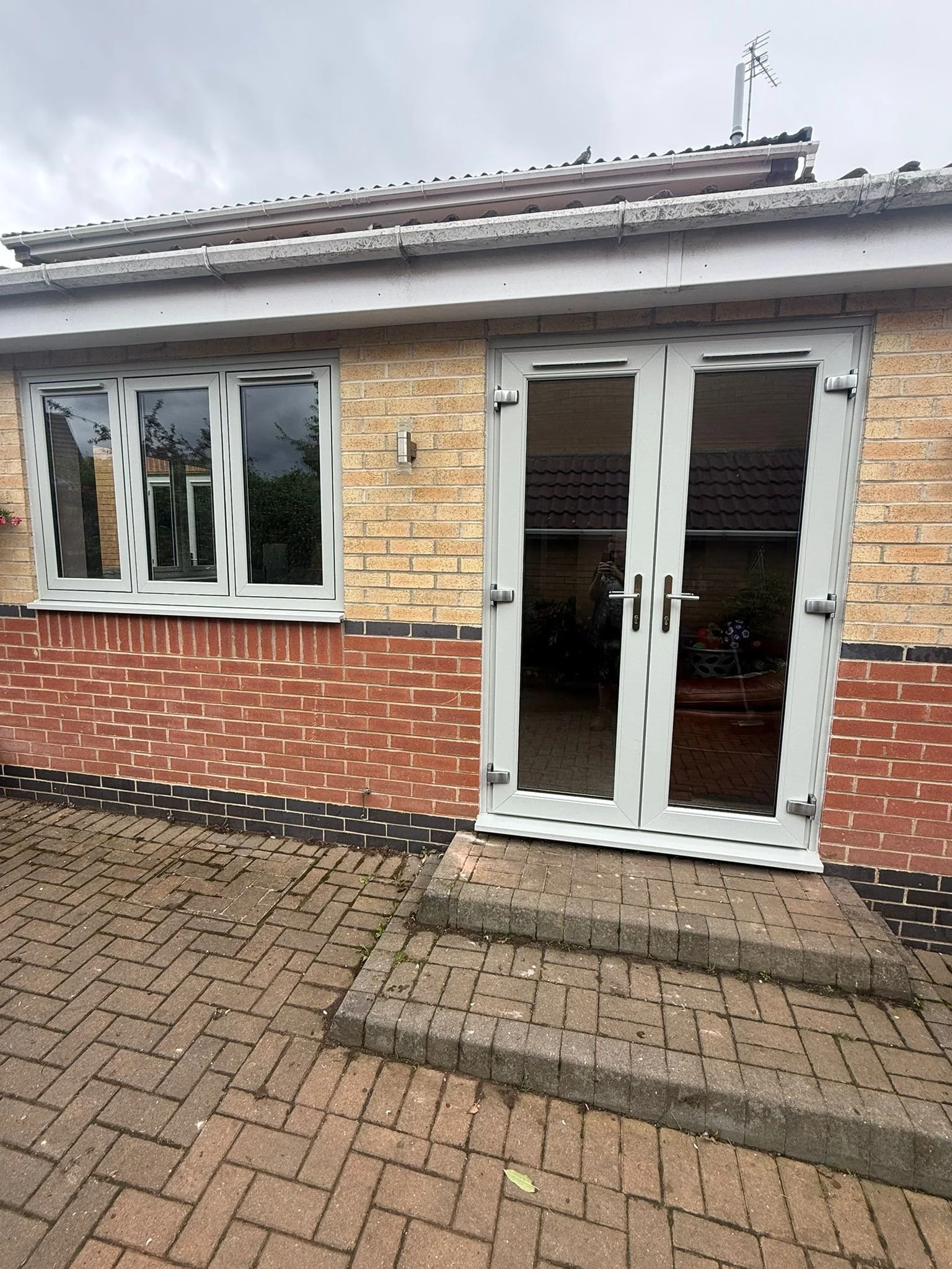 View of a house's exterior showing a brick and brick veneer wall, a white-framed glass door with a handle, and a window with white framing. The paving is made of brick pavers, with a concrete step outside the door.