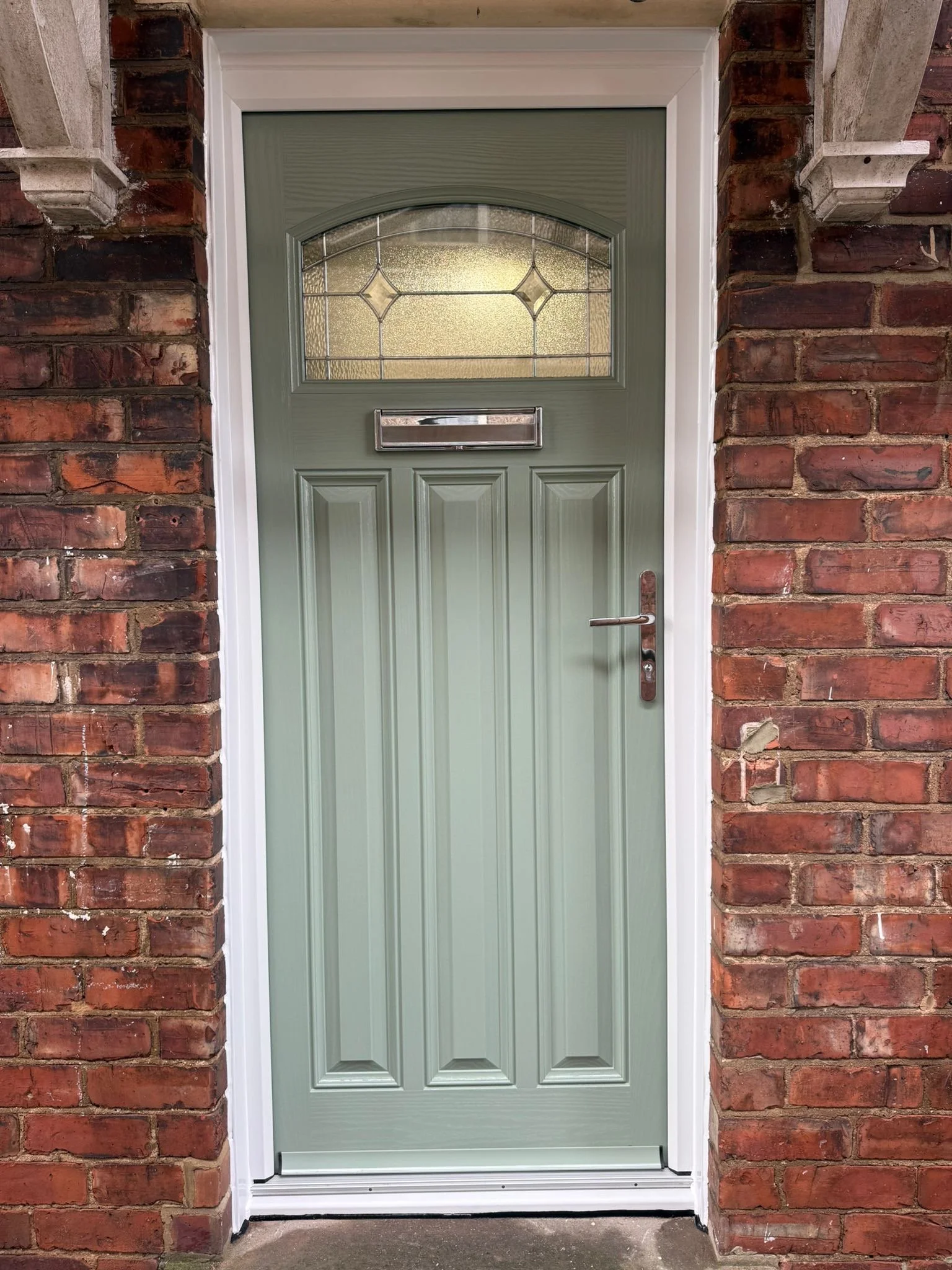 A green front door with decorative glass window, mail slot, and silver handle, surrounded by a white frame and brick walls.