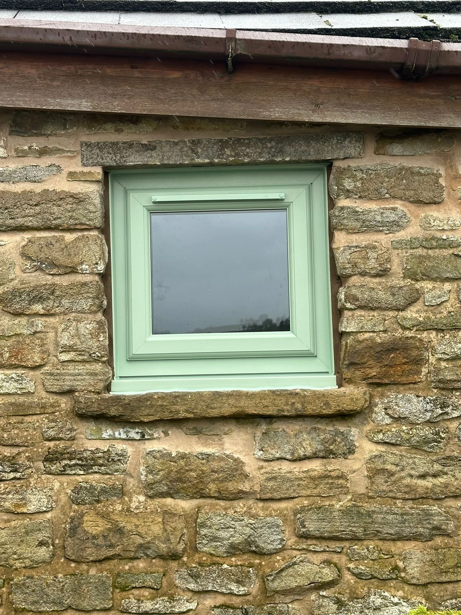 A small rectangular window set into an aged stone wall with irregularly shaped stones and mortar. The window has a pale green frame and a reflective glass pane. Above it is a stone lintel, and just above that, a wooden beam with a gutter attached.