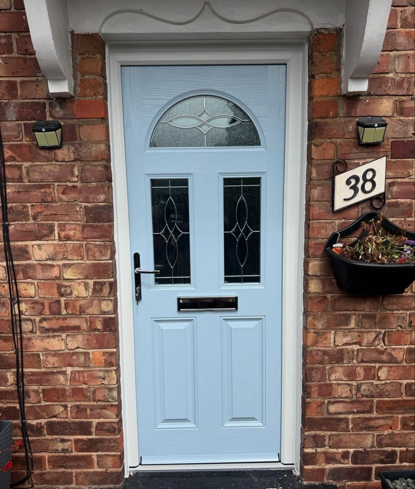 Blue front door with decorative glass panels, surrounded by red brick wall, with house number 38 on a white sign to the right, and a hanging flower basket below the sign. Two black outdoor lights are mounted on the brick wall, one on each side of the