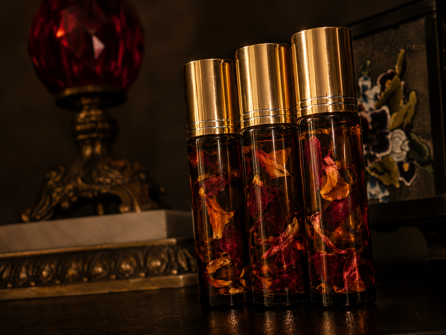 Three glass bottles with gold caps containing dried flower petals, placed on a dark surface in front of ornate decorative objects.