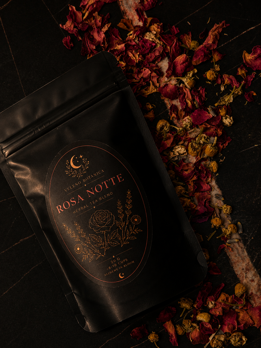 A black package labeled 'Rosa Notte' herbal tea blend lying on a dark surface, with dried red and yellow flower petals and herbs scattered around it.