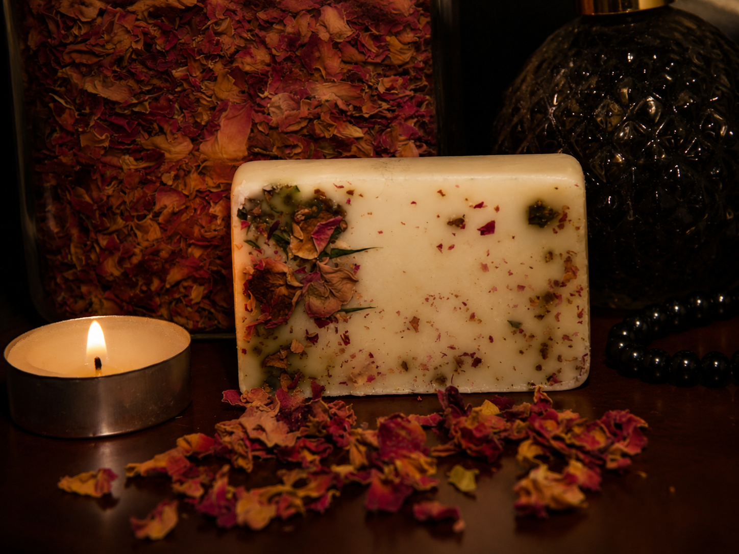 A white rectangular soap bar embedded with dried rose petals and herbs, surrounded by scattered dried rose petals, a lit tea light candle, a black decorative object, and a container filled with more dried rose petals.