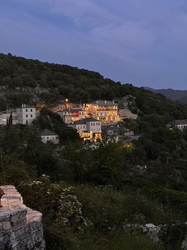 Hillside village with houses illuminated at dusk, surrounded by green trees and a mountain backdrop.