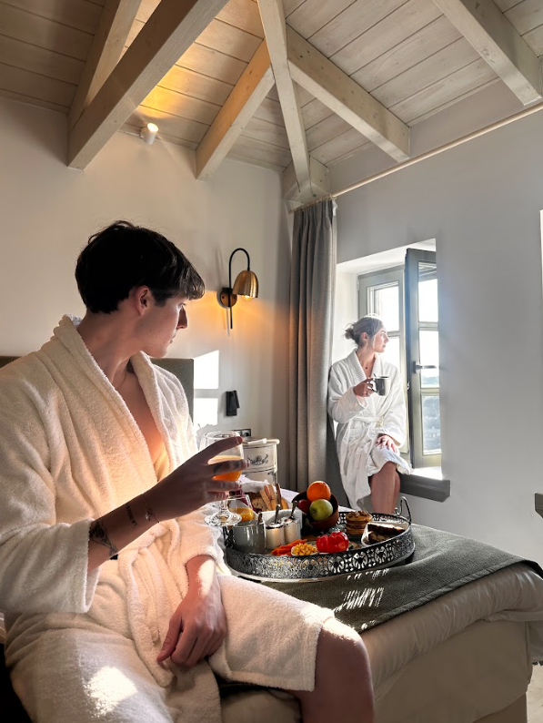 Two women in bathrobes enjoying breakfast in a bright, cozy room with a wooden ceiling. One is sitting on the bed holding a glass, the other is sitting on the windowsill with a cup, looking outside.