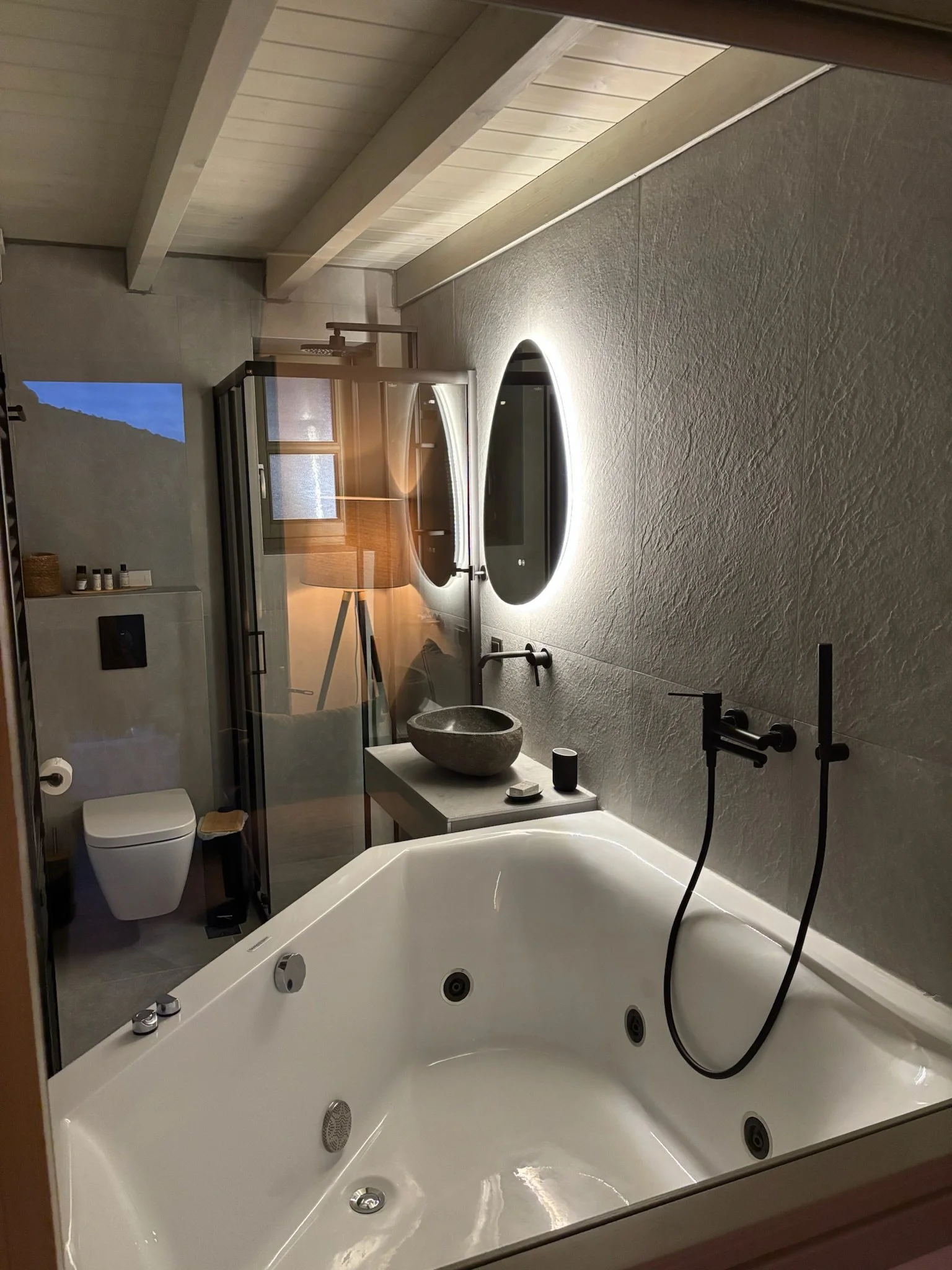 Bathroom with a large whirlpool bathtub, gray textured wall, window, and shower area separated by a glass partition. Includes a toilet, small table, and various toiletries.