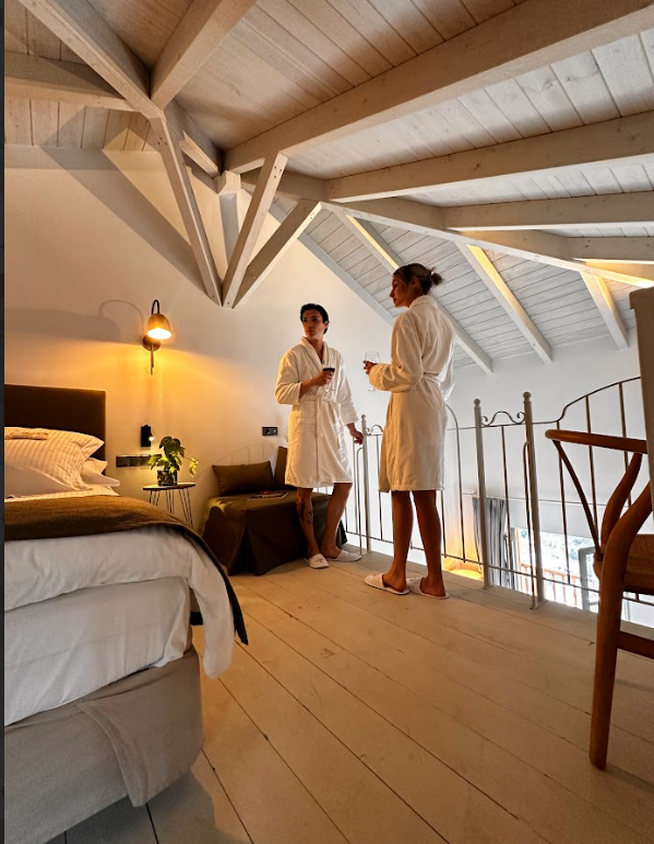 Two women in white bathrobes standing and talking in a cozy, well-lit attic bedroom with a slanted ceiling and exposed wooden beams, near a railing overlooking a lower level.