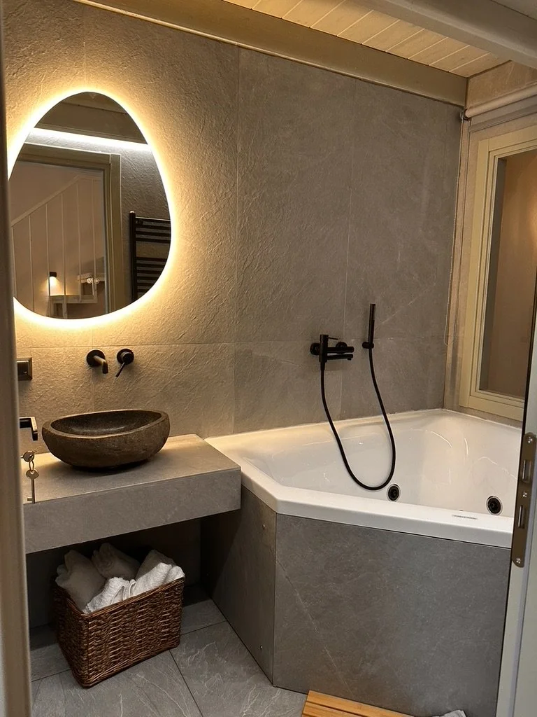 Bathroom with a wall-mounted mirror with backlighting, a stone vessel sink on a concrete counter, a basket with towels underneath, a corner bathtub, and a window on the right.