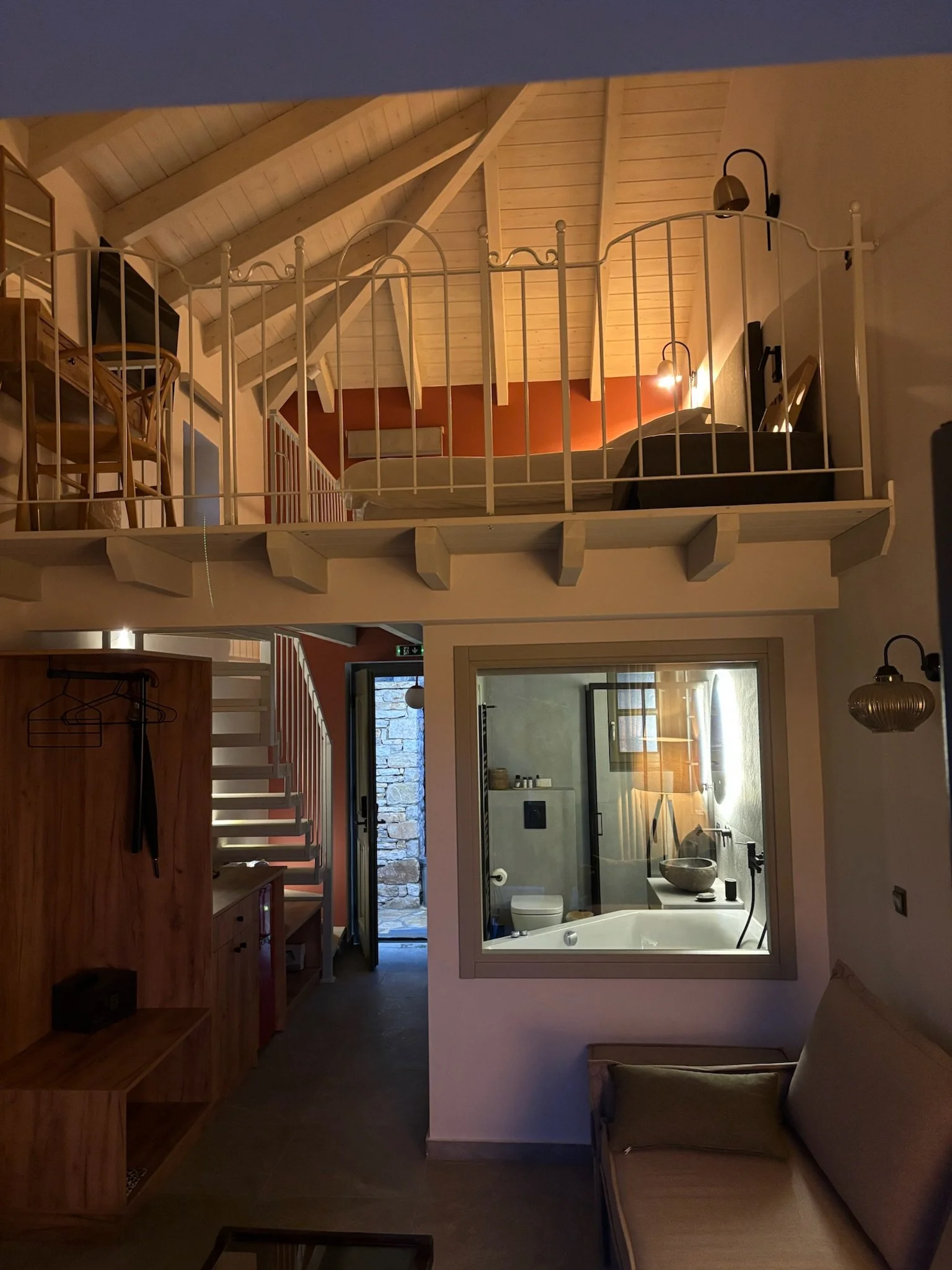 Interior view of a cozy room with a loft area, wooden ceiling, and a bathroom visible through a glass door. The room is decorated with warm lighting and modern furniture.