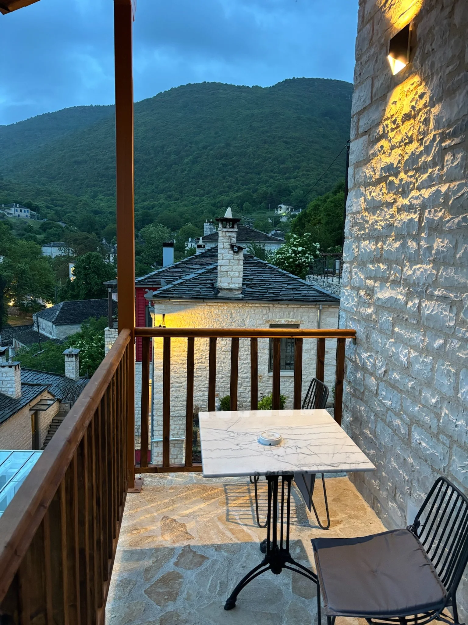 A small balcony with a marble table and black chairs overlooking a neighborhood with houses and green hills in the background during dusk.
