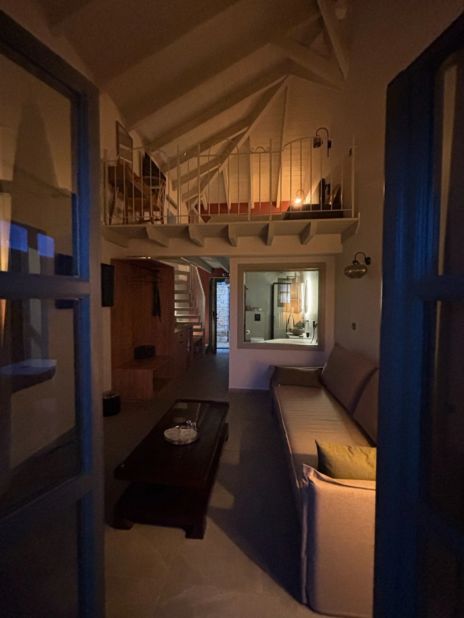 A cozy living room lit by warm, ambient light, with a sofa against the wall, a coffee table with a decorative item, a small table with a lamp, an open hallway leading to other rooms, and a loft with a chair and stairs visible.