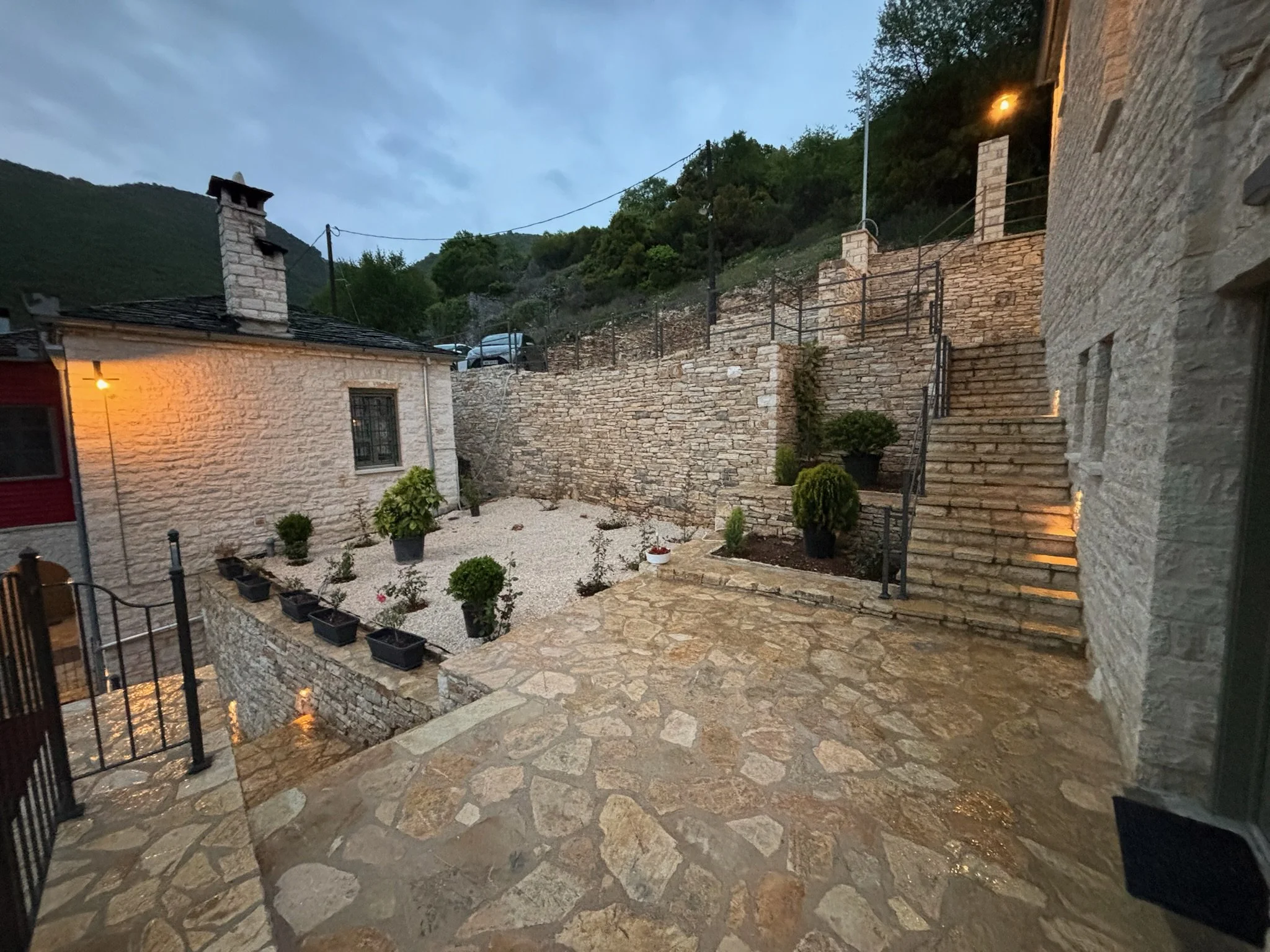 Exterior view of a stone courtyard at dusk, featuring stone stairs, potted plants, a stone wall, and a hill with trees in the background.