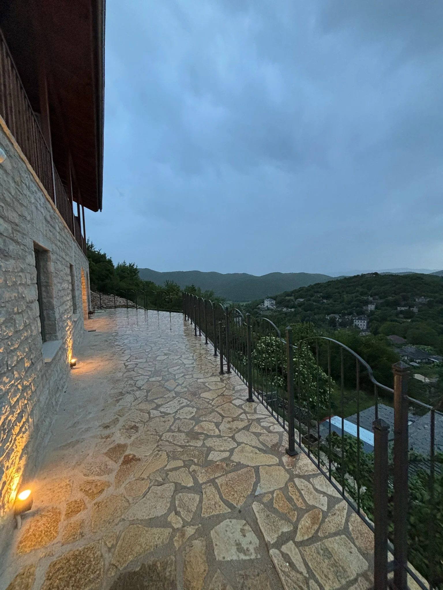 Terrace with stone paving, lit by warm lighting, overlooking hills and a small town at dusk.