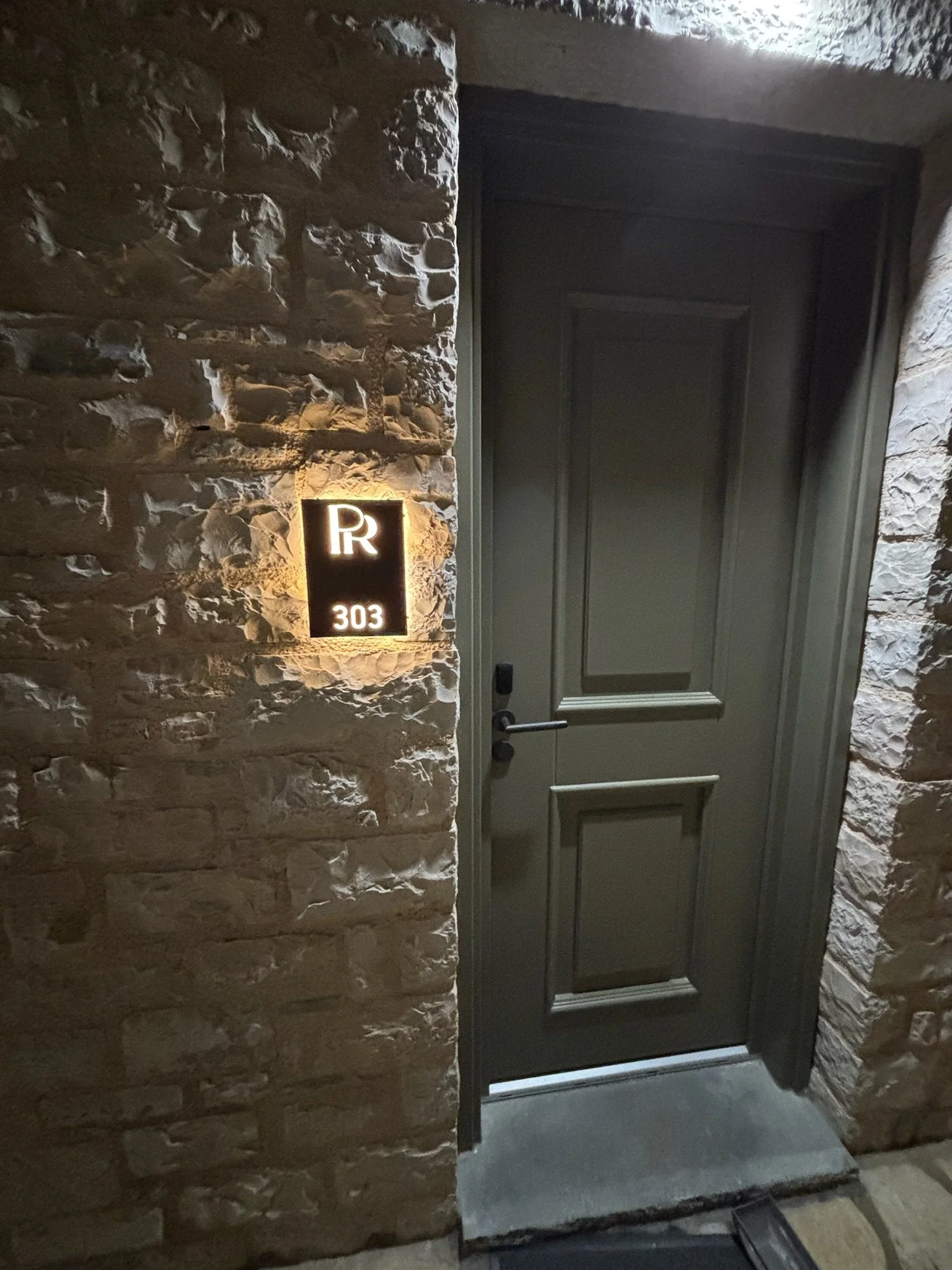 A stone wall with a black door and a illuminated sign displaying the letter 'R' and the number '303' next to it.