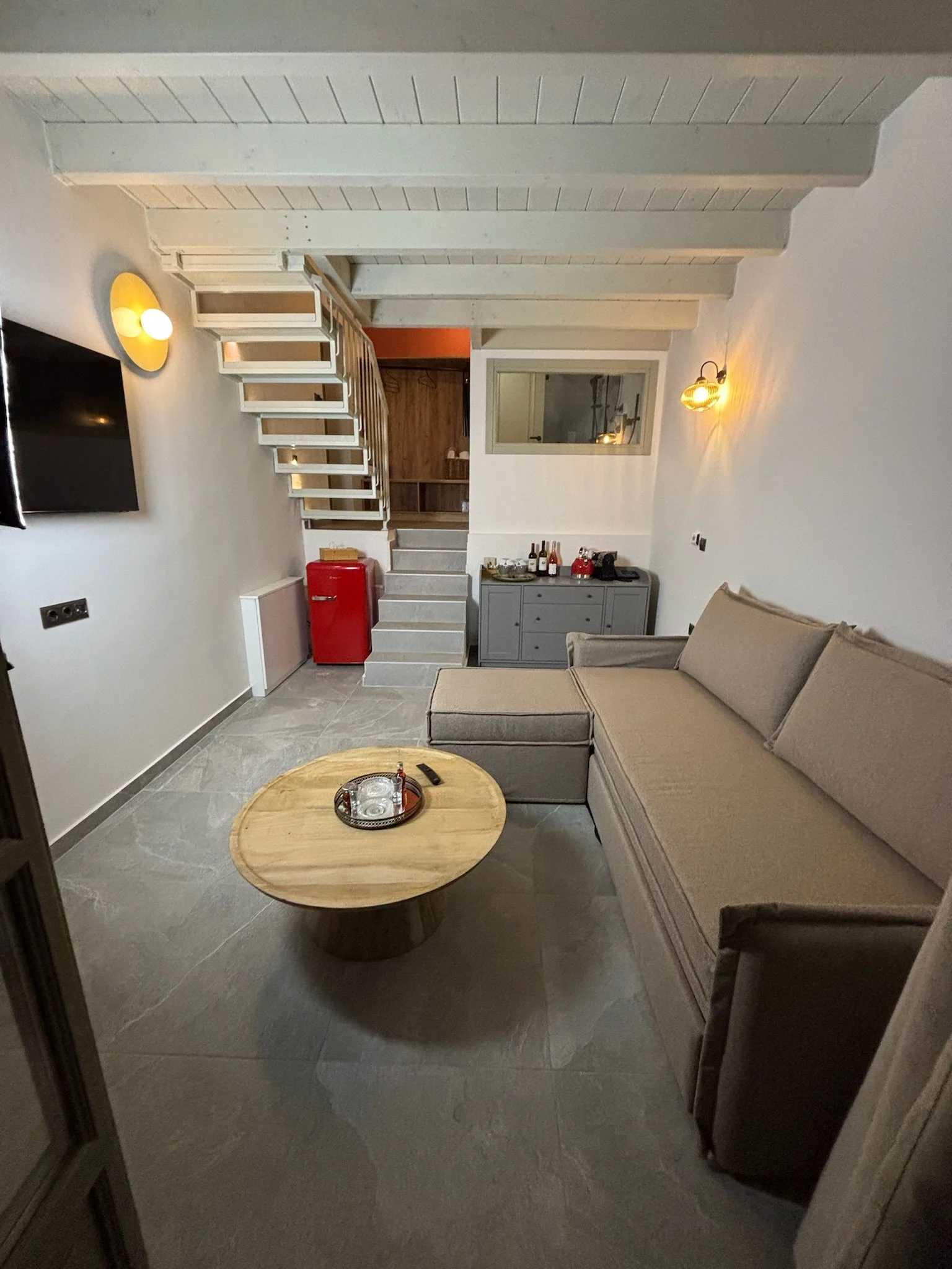 Living room with a beige sofa, a round wooden coffee table, a gray sideboard decorated with wine bottles and glasses, wall-mounted lights, and a staircase leading to an upper floor.