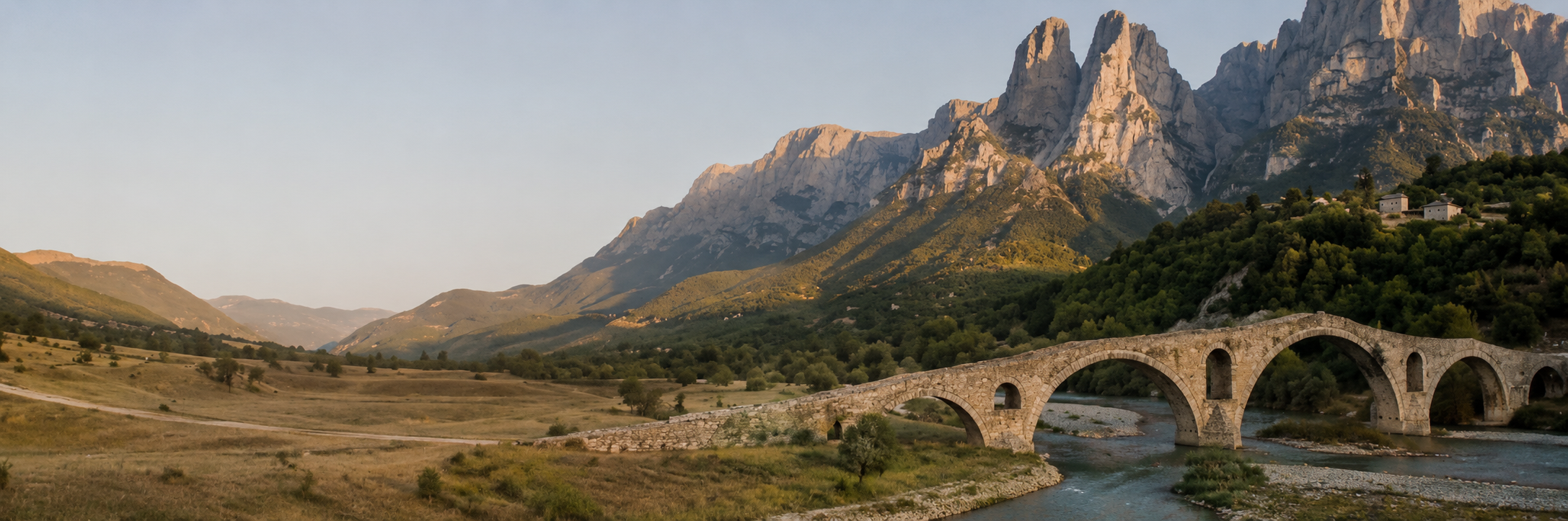 Scenic landscape featuring a stone bridge over a river with green hills and tall mountains in the background during sunset.