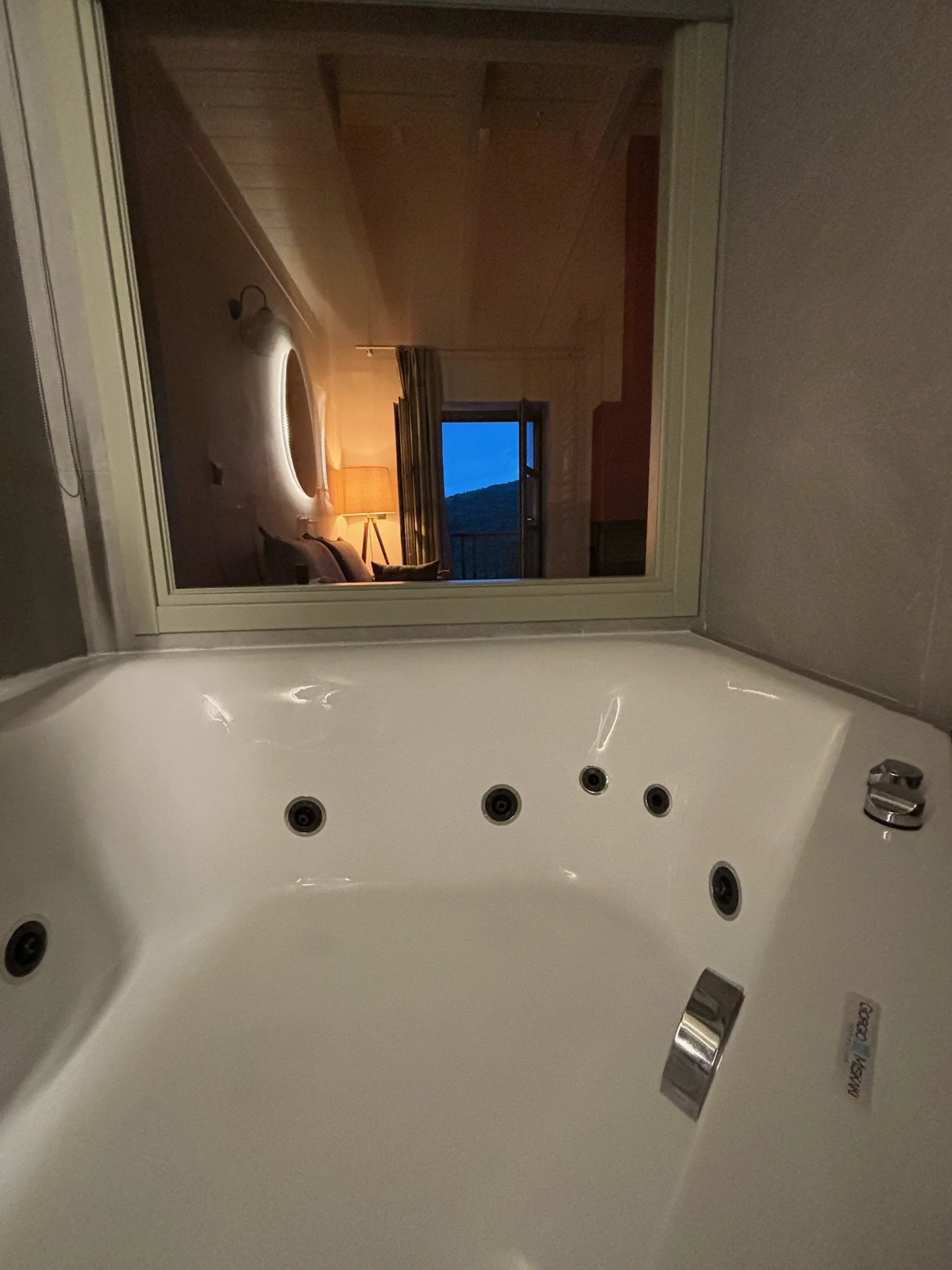 A large white whirlpool bathtub with multiple jets and a chrome faucet, viewed from inside the tub looking outward at a mirror reflecting the room and a window.