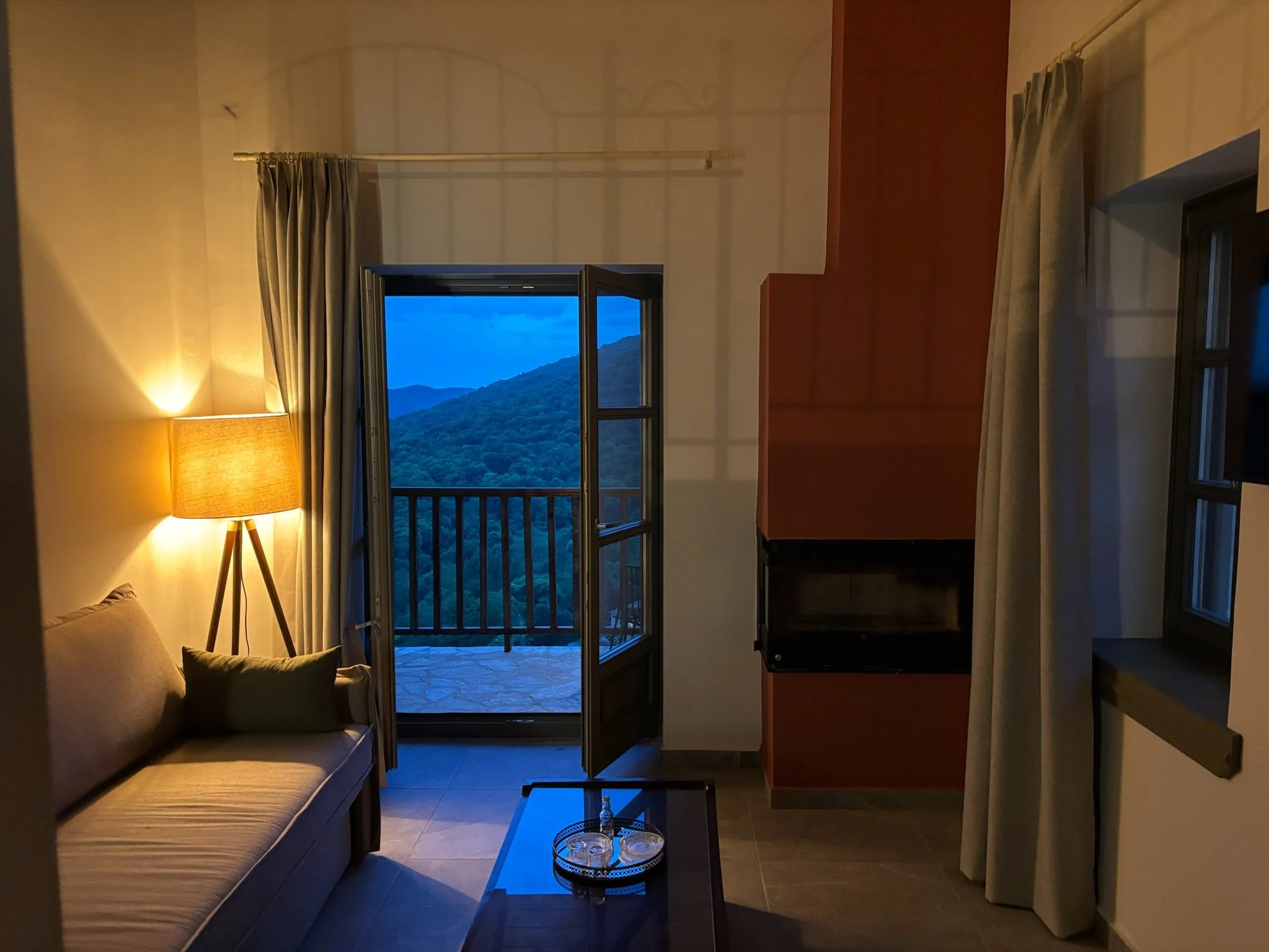 Interior of a room at dusk with a beige sofa, a floor lamp turned on, a small black table with two glasses, a balcony door open showing a mountain landscape, and curtains on either side.