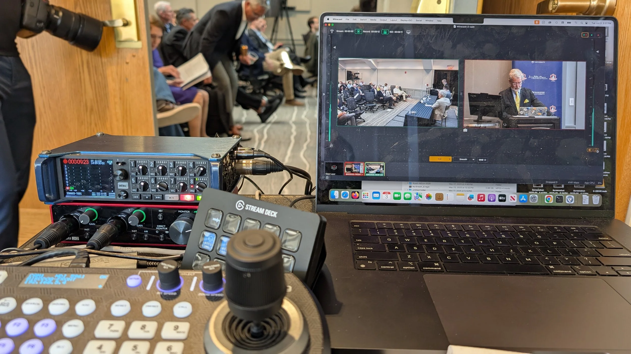 View of a camera and audio equipment setup on a table with a laptop displaying a video conferencing platform. In the background, a group of people seated in a room, some reading, appears to be attending a presentation or meeting.