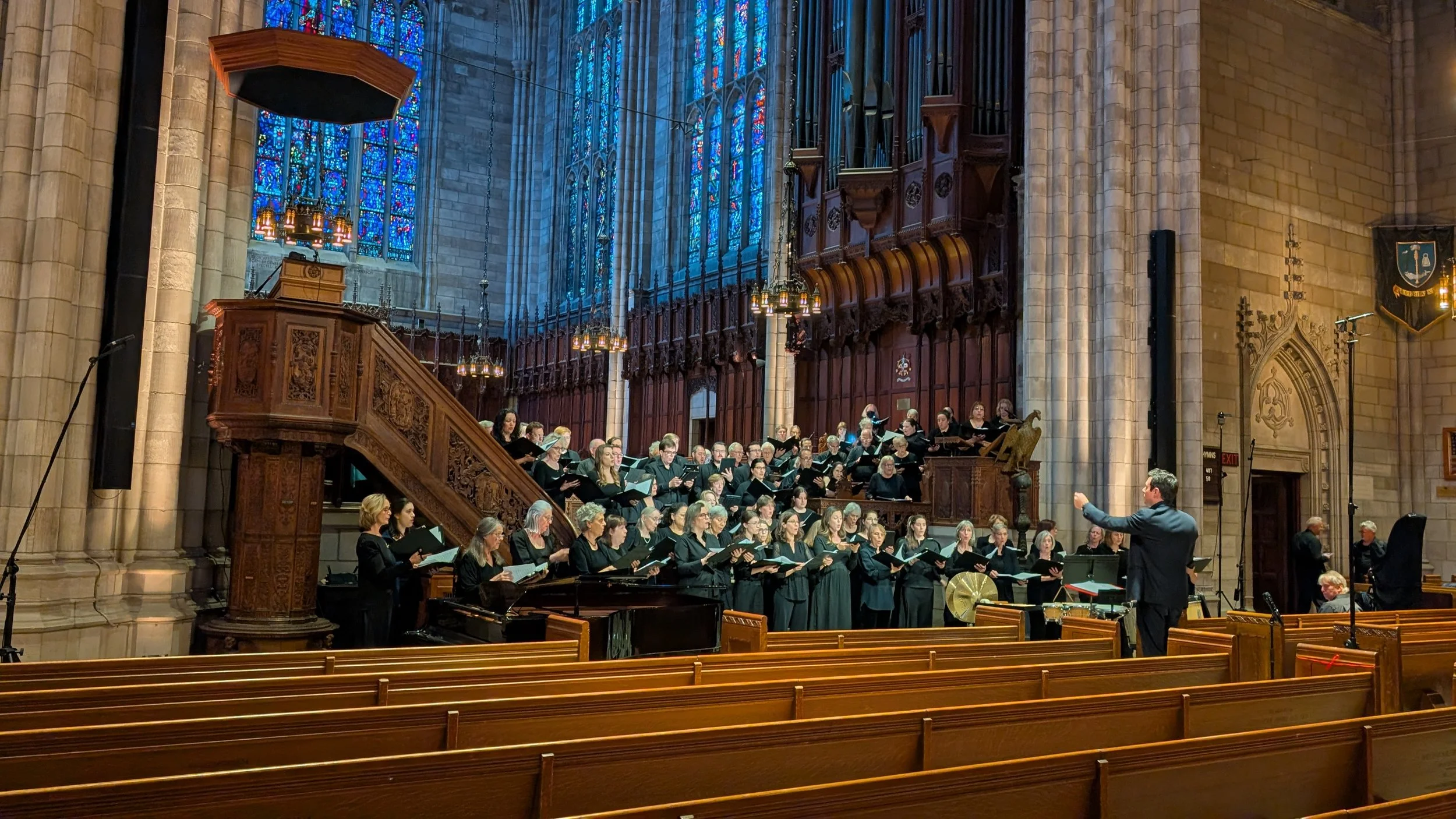 A large choir performing inside a grand church with tall stained glass windows and intricate woodwork, led by a conductor.