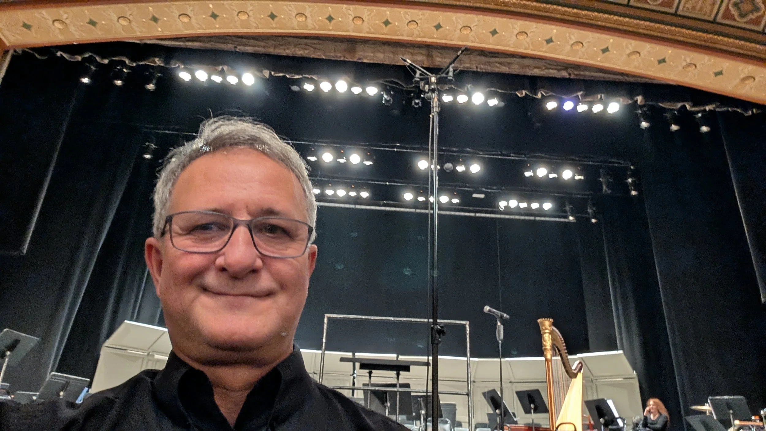 Close-up selfie of a man with glasses smiling, standing on a stage inside a concert hall with a black backdrop, stage lights, music stands, and musical instruments in the background.