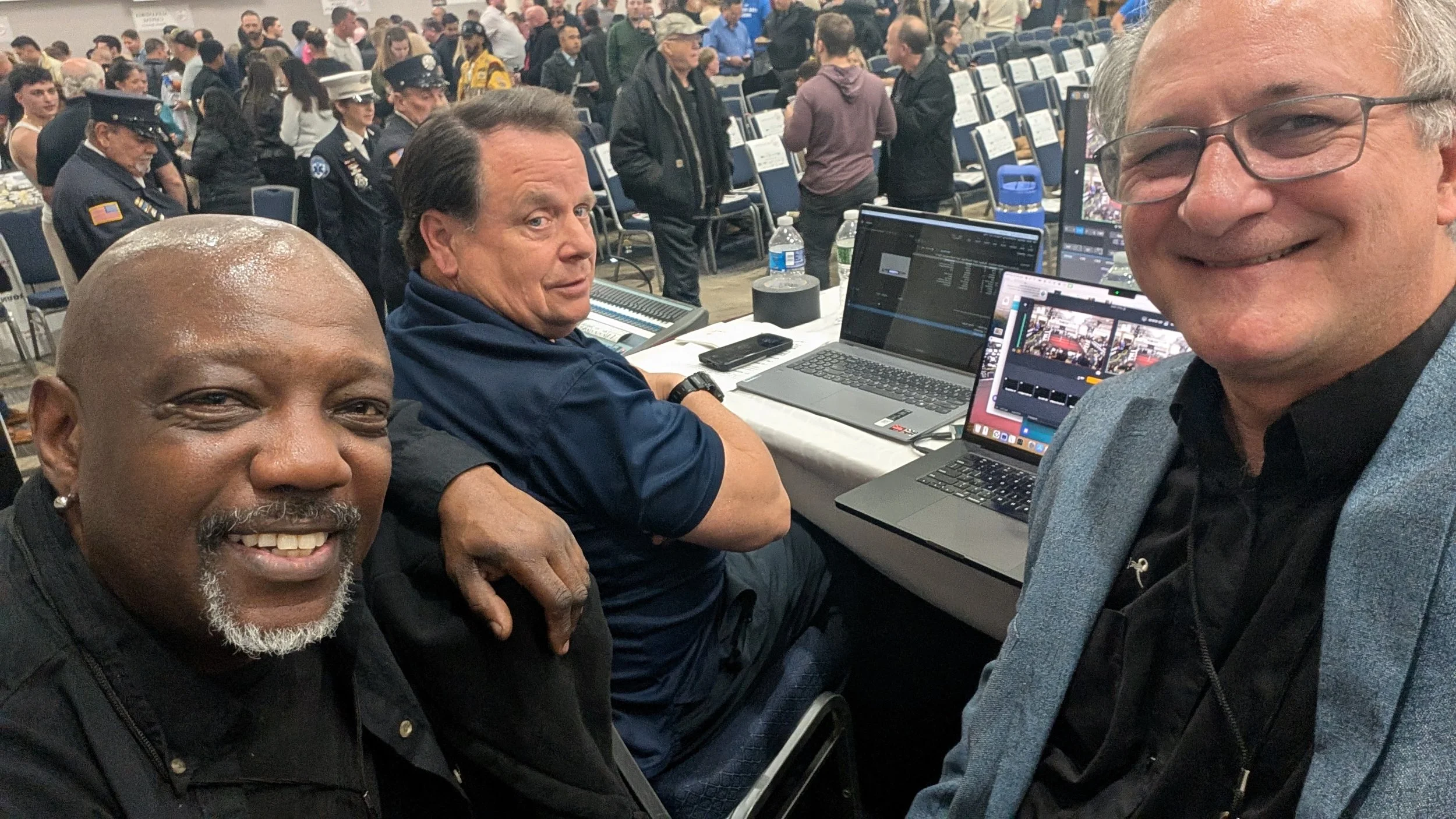 Three men taking a selfie at an indoor event with many people, some in uniform, and rows of empty chairs in the background. The man on the left has a beard and a smile, the man in the middle is looking at the camera with a neutral expression, and the man on the right is smiling broadly.