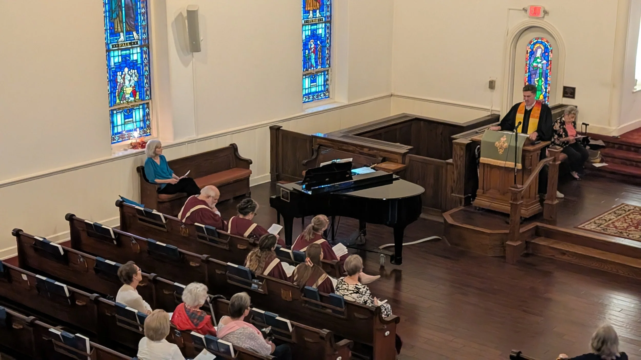 Inside a church with stained glass windows, a congregation seated in pews, a woman sitting on a bench, a black grand piano, and a man and woman at the pulpit.