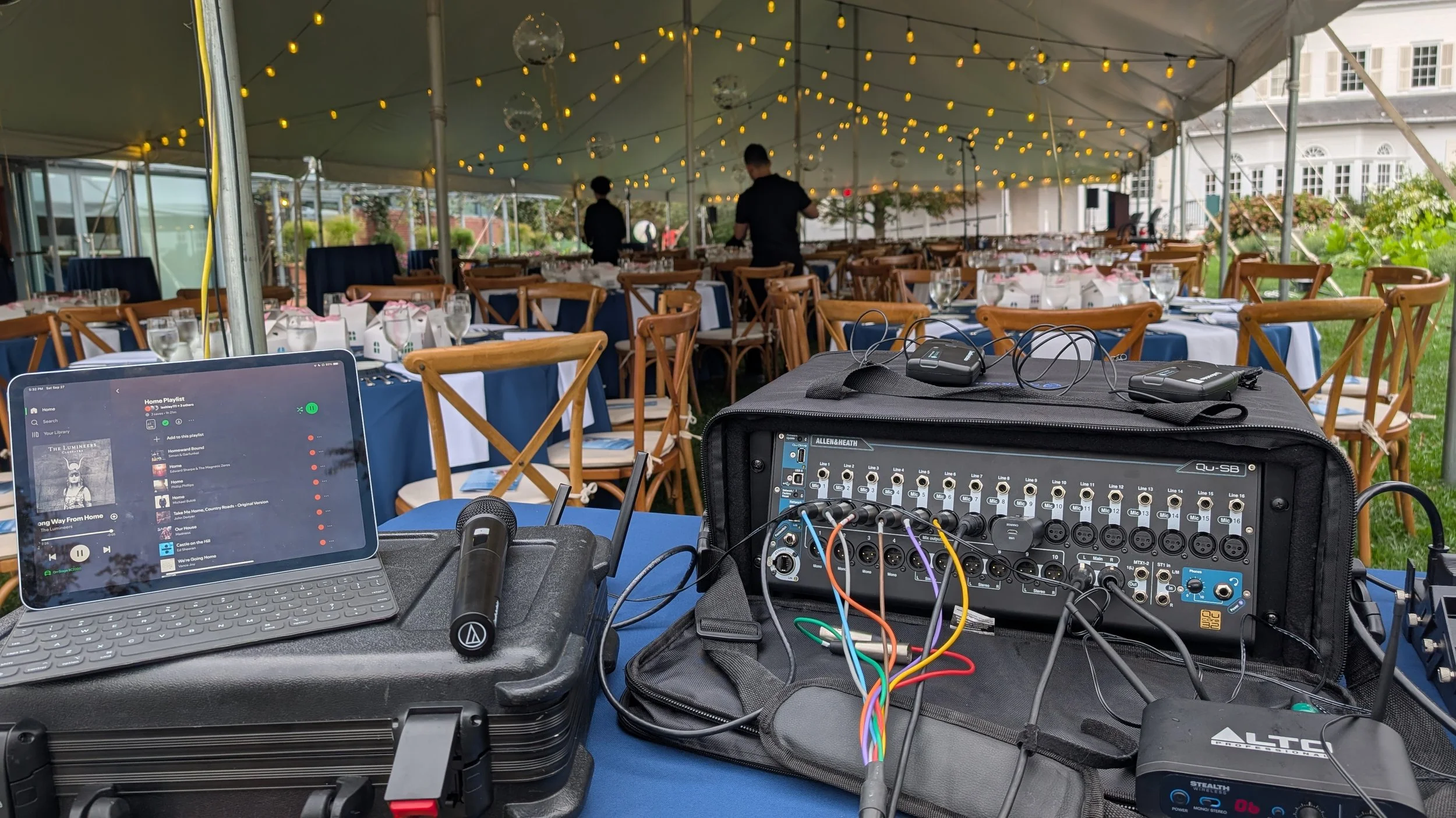 Audio equipment setup under a tent at an outdoor event, with a laptop playing music, a sound mixer, and two wireless microphone packs, surrounded by tables and chairs with table settings and string lights.