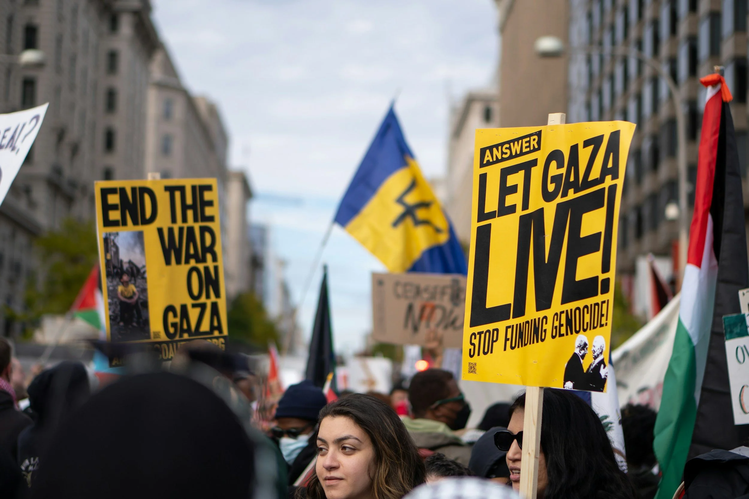 A crowd of protesters holding signs, including one that reads 'Let Gaza Live', with a flag in the background, on a city street with buildings.