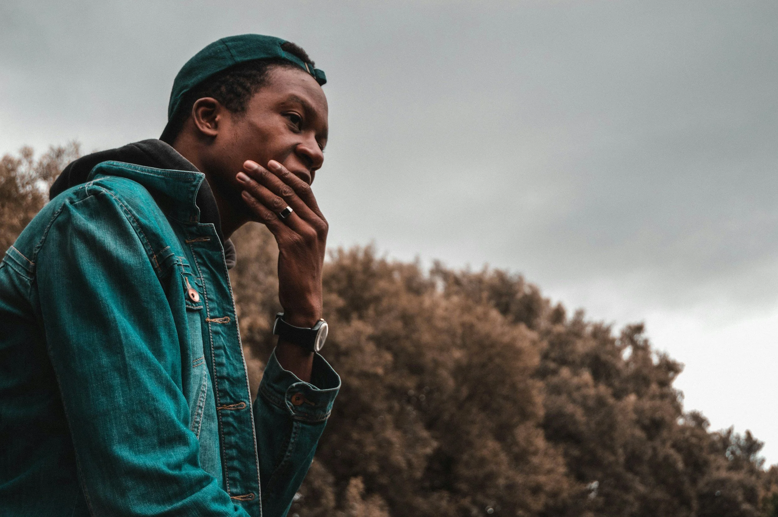 A young man wearing a backwards baseball cap and a denim jacket, holding his chin thoughtfully with his left hand, seen outdoors on a cloudy day with trees in the background.