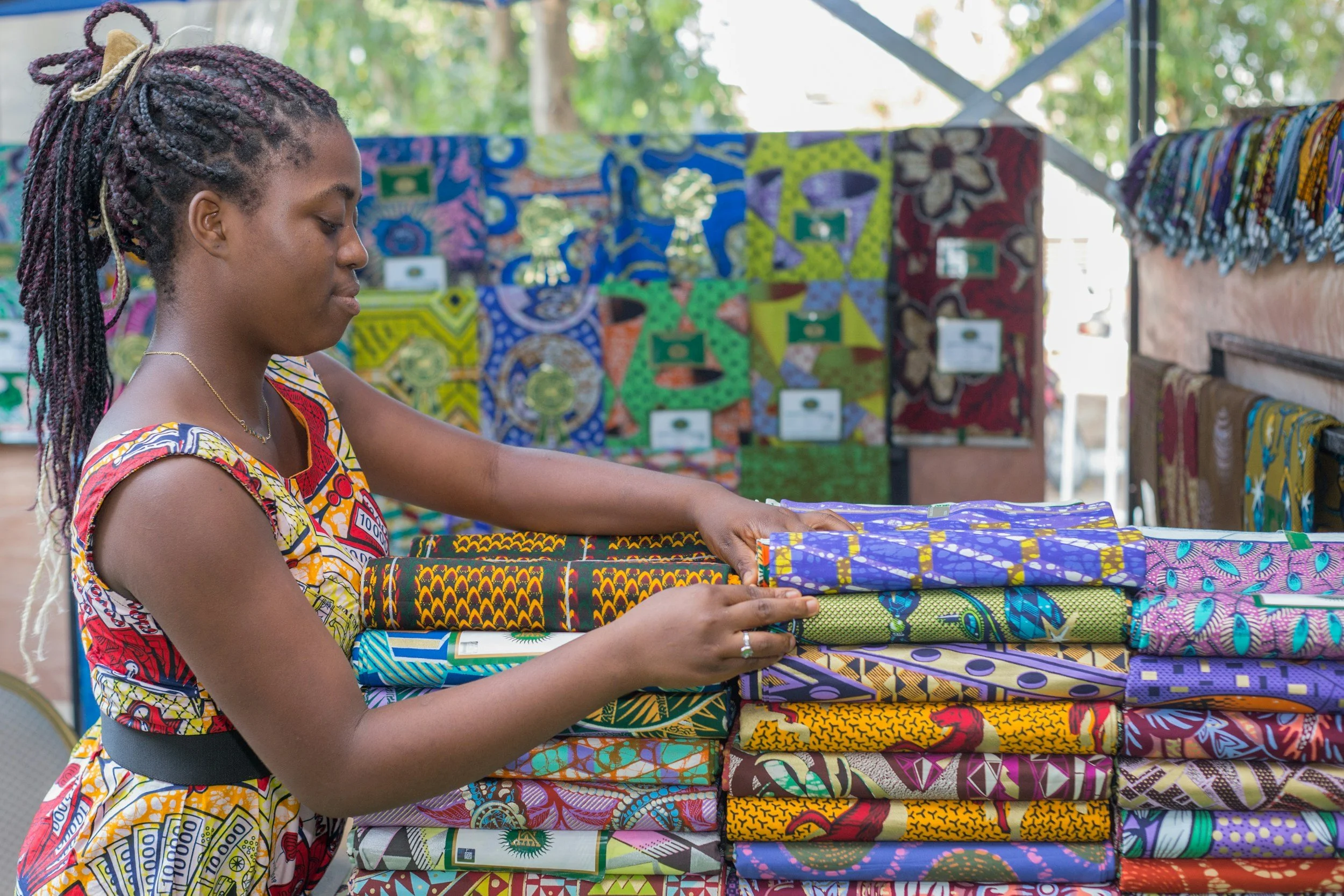 A woman with braids and colorful clothing organizing vibrant fabric at a stall, with more fabric and a blurred background of trees.