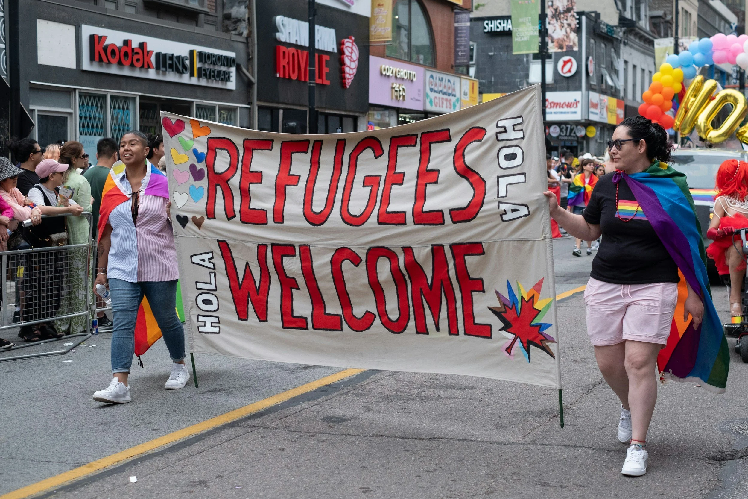 People marching in a pride parade holding a large banner that reads 'Refugees Welcome' decorated with colorful hearts and stars, on a city street lined with storefronts and balloons.