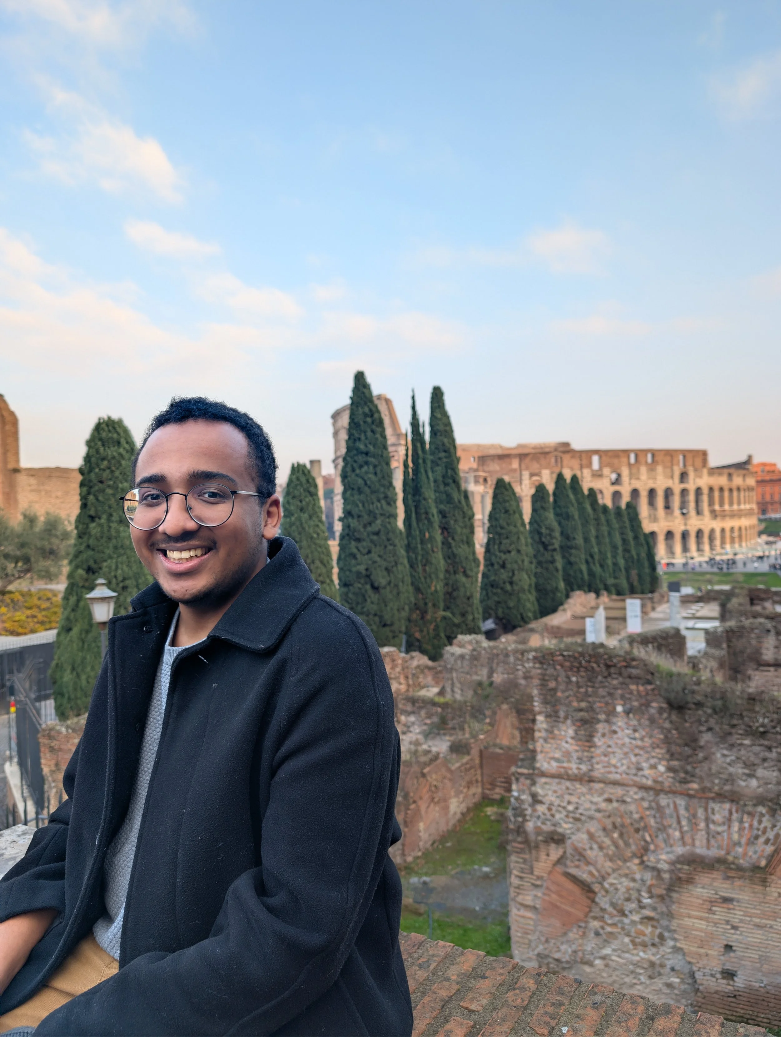 A young man with glasses and a smile sitting outdoors in front of ancient ruins and tall cypress trees, with a bright sky overhead.