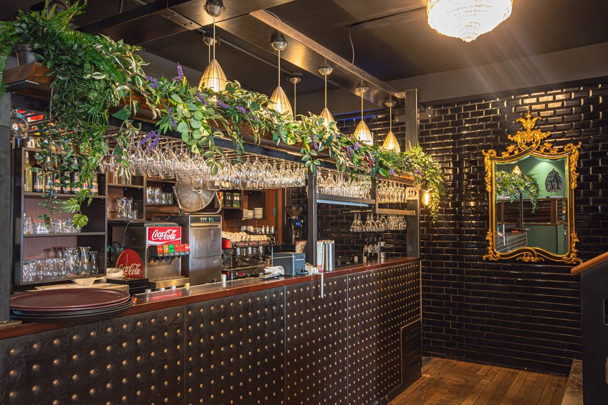 Un comptoir de bar dans un restaurant ou un café avec des étagères de verres, des plantes suspendues, un miroir doré, et un mur noir en briques.