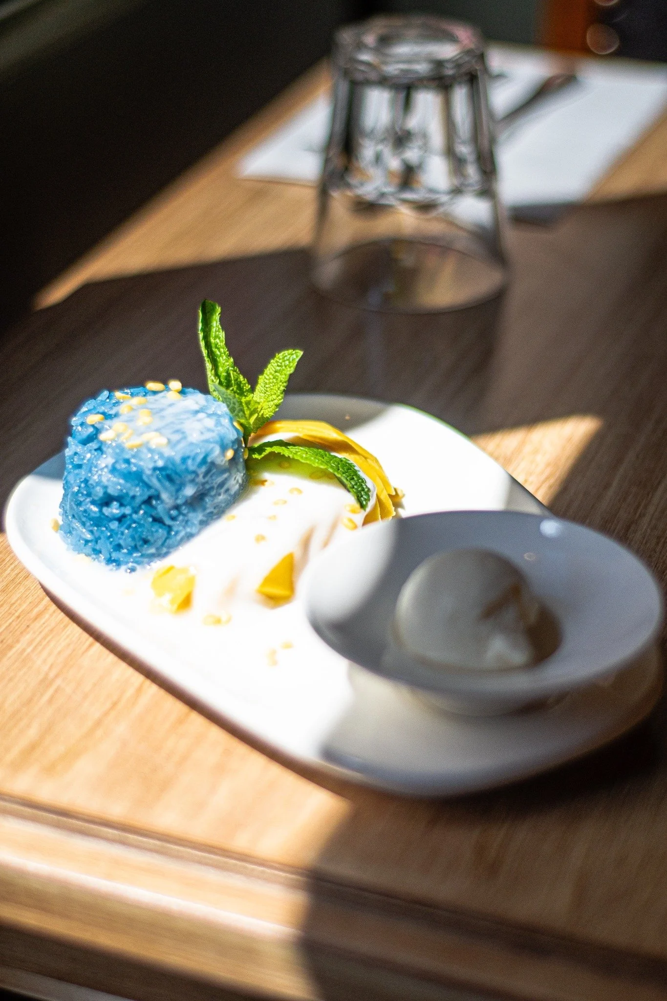 Un plat de dessert avec une boule bleue, des feuilles de menthe, et une rondelle de citron, accompagné d'une cuillère avec une portion de crème ou de pudding.