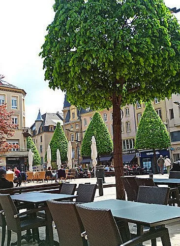Une place urbaine avec des tables et chaises d'extérieur, entourée d'arbres et de bâtiments historiques en arrière-plan, par une journée ensoleillée.