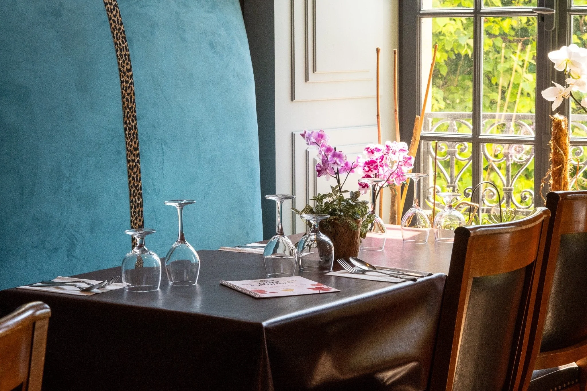 Une table à manger décorée avec des verres, des couverts, une nappe noire et un menu, dans une salle avec une grande fenetre laissant entrer la lumière, et des plantes et fleurs dans le décor.
