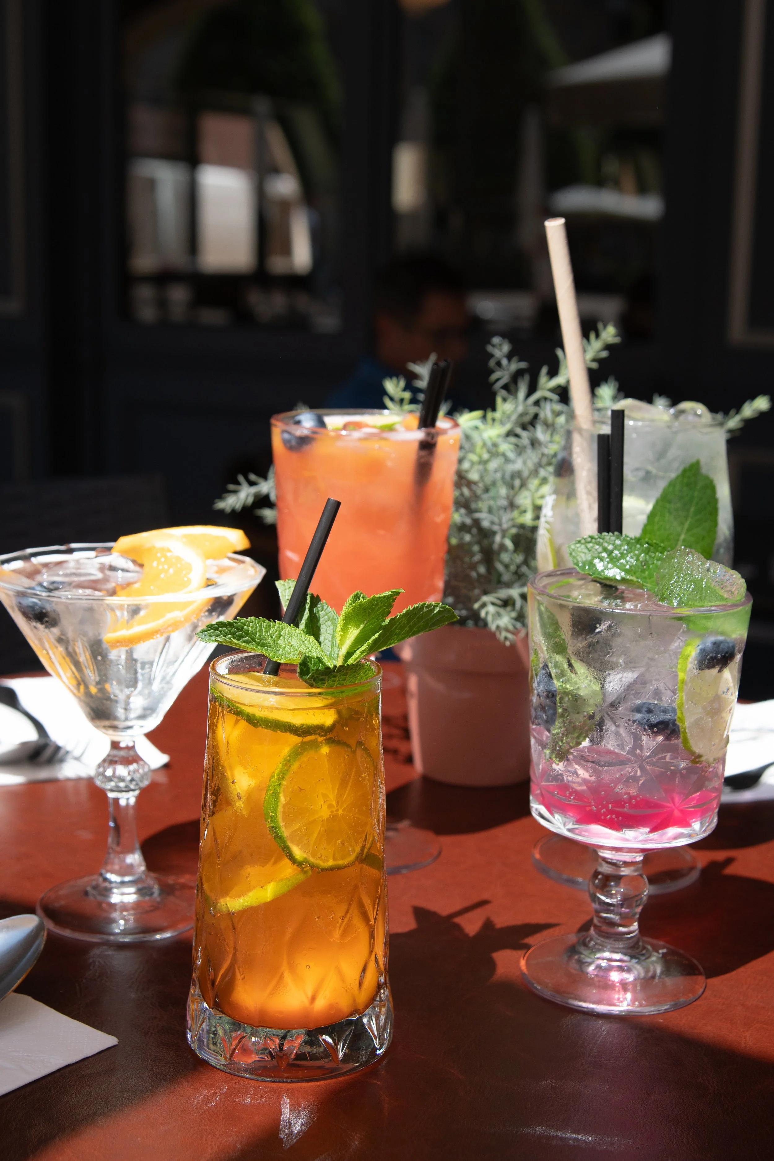 Plusieurs cocktails colorés avec des herbes et des fruits, sur une table en bois. En arrière-plan, une personne floue dans un environnement intérieur, probablement un café ou un restaurant.