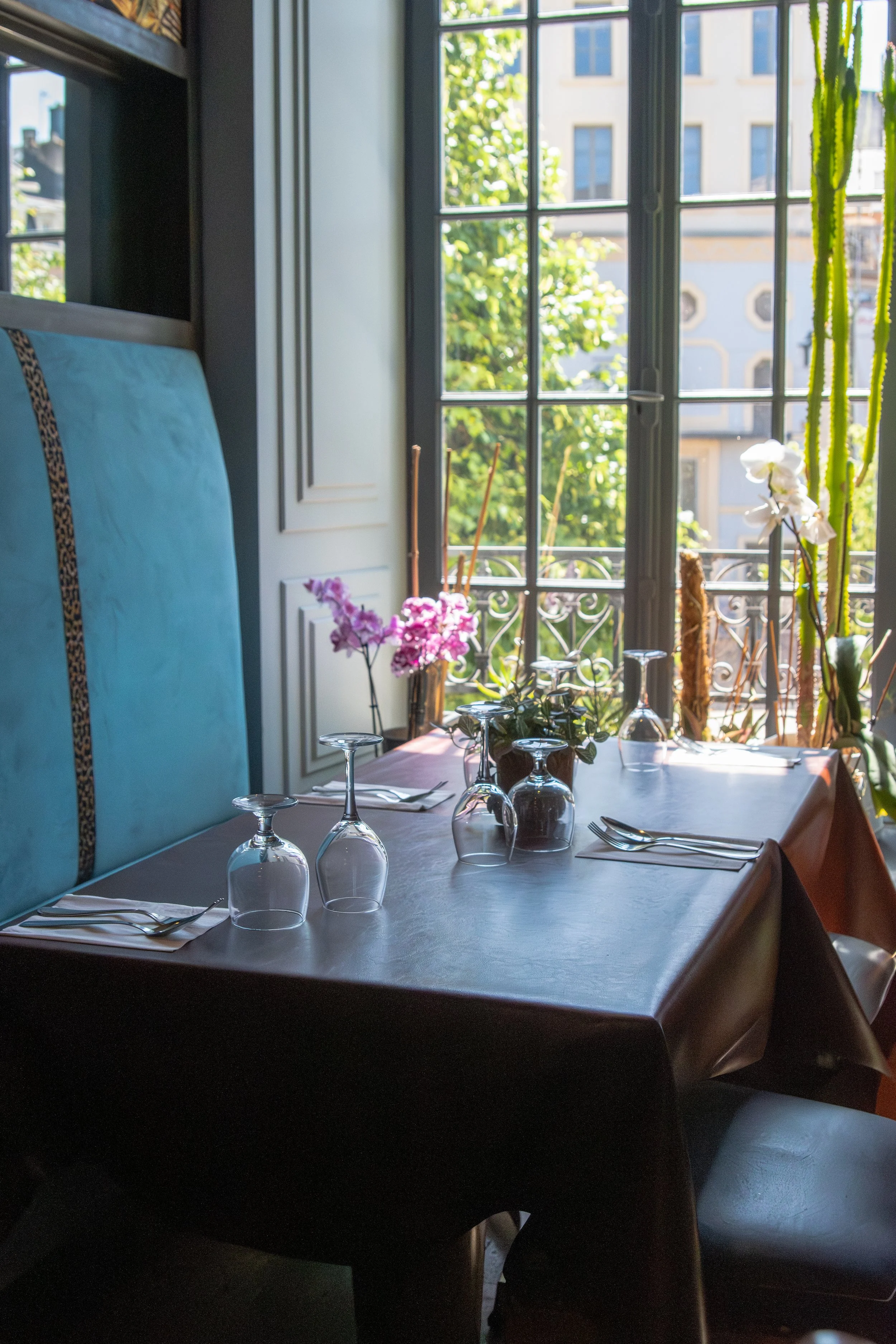 Une salle à manger avec une table dressée, des verres à vin inversés, des couverts, des fleurs en pot sur la table, dans une pièce lumineuse avec de grandes fenêtres laissant entrer la lumière naturelle, vue sur des bâtiments et des arbres.